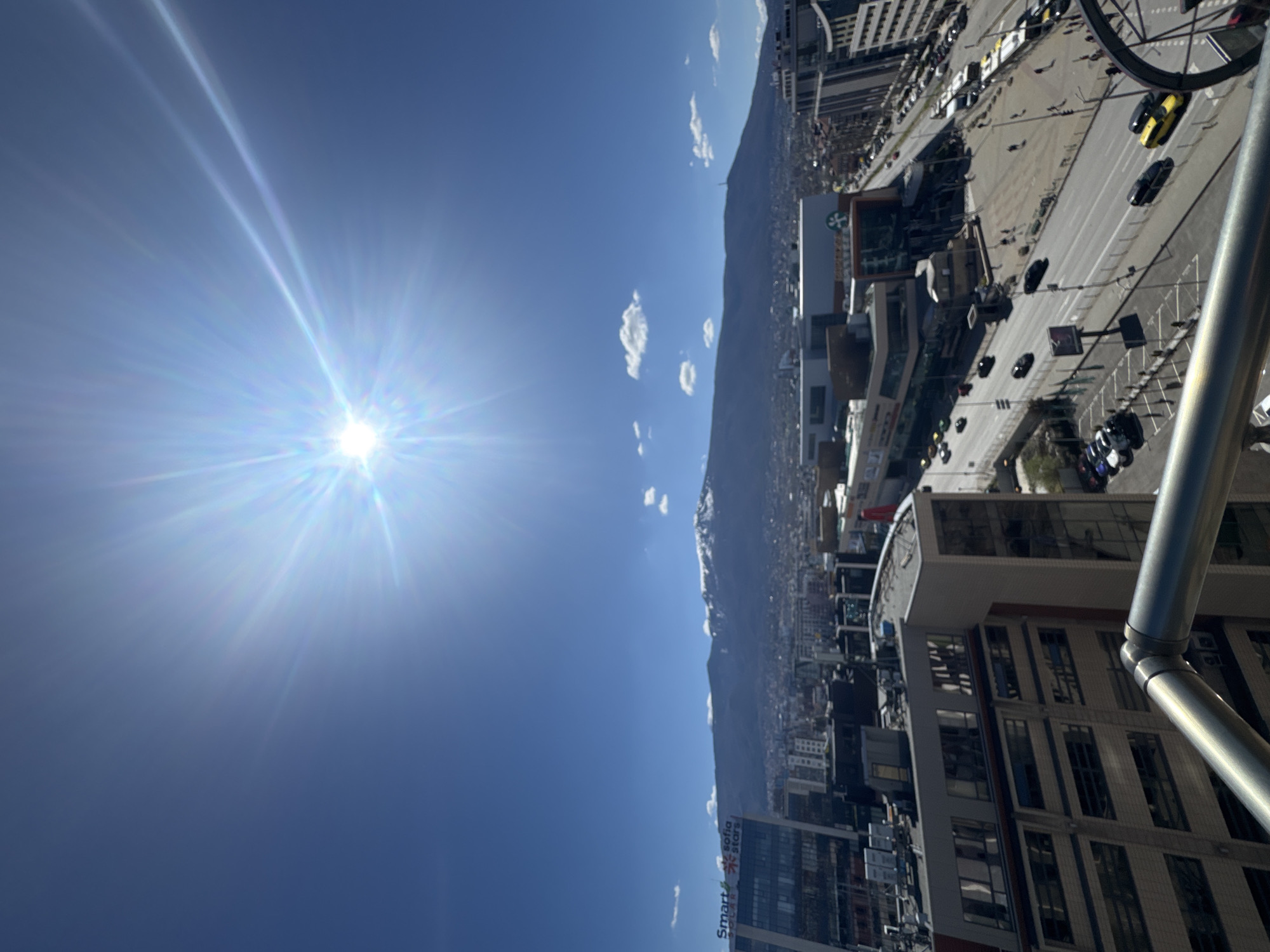 The image shows a view of Sofia, Bulgaria, on a sunny day. The sun is visible in the blue sky with a few scattered clouds. In the distance, there is a mountain range. Below the mountain, the city stretches out, with buildings and streets visible. Cars are driving on the roads. There's a large structure of scaffolding on the right. "Smart Sofia Stars" are written on a building.