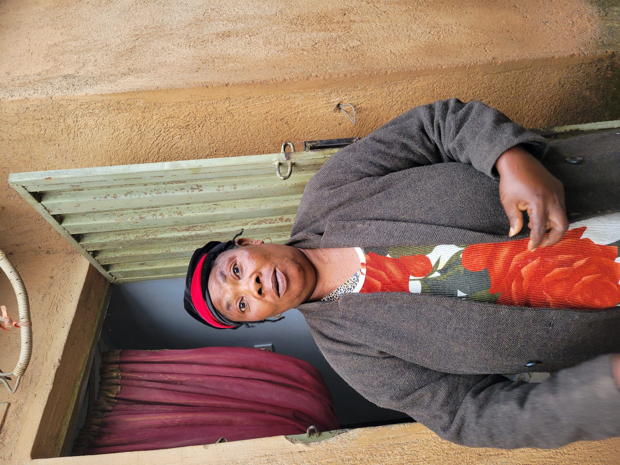 An outdoor, medium shot captures a Black woman standing in a doorway in an unknown city. She is positioned slightly off-center to the left and faces the camera directly, her eyes wide and her mouth slightly open as if she is speaking. She wears a dark brown textured jacket over a dress with a bold floral print featuring large red and green flowers. On her head is a black and red patterned cap. Her skin is dark, and she has prominent facial features, including a round face and dark eyes. She is wearing a gold hoop earring in her visible ear. The doorway has a light brown wooden frame and a weathered, light green metal gate that is partially open. Behind her, a dark red curtain is visible. The wall to her right is a rough, textured light brown. The overall lighting suggests an overcast day.
