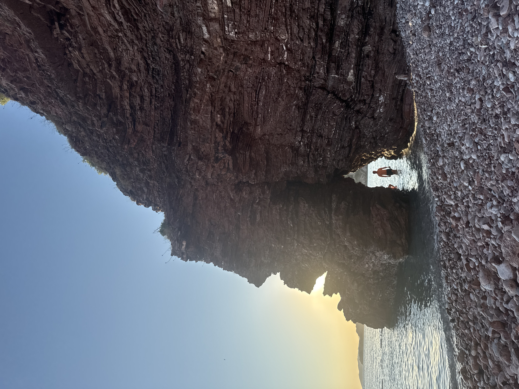 In Sveti Stefan, Montenegro, a person stands in shallow water near a rocky archway. The sun is setting, casting a warm glow over the scene. The beach is covered in pebbles, and the water is calm and clear. The rock formations are a deep reddish-brown, with visible layers and textures. The sky is a clear, pale blue.