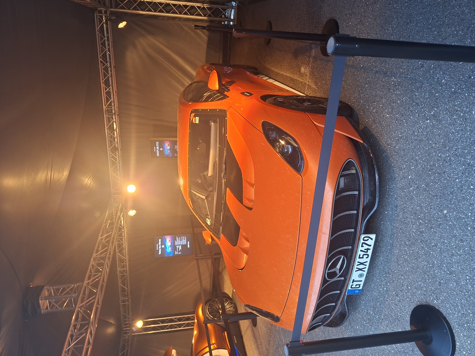 An orange Mercedes-AMG GT XX concept car is displayed under a tent with industrial lighting and truss structures. The car is parked on an asphalt surface and is cordoned off by black stanchions and grey velvet ropes. A license plate reading "GT XX 5479" is affixed to the front of the car. In the background, a digital display shows information about the "Vision One-Eleven." To the left, another orange concept car is partially visible. The image appears to be taken during an event at the Mercedes-Benz plant in Rastatt, Germany, specifically within the facility's event or exhibition space. There are no people visible in the frame.