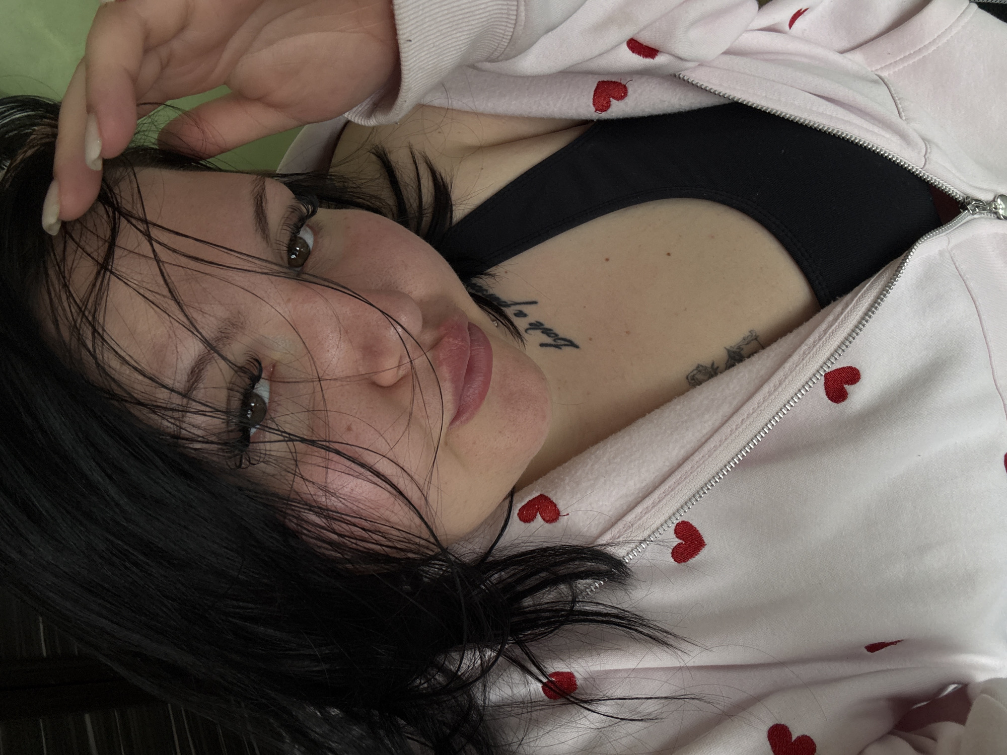 A young woman lies supine, partially visible. She is wearing a light pink jacket with red hearts, partially unzipped, revealing a black bra or tank top underneath. Her dark hair is disheveled, with some strands falling across her face. She is touching her forehead with her hand, her gaze directed toward the camera. She has a tattoo on her collarbone, visible on the upper right part of her chest. The background is indistinct, and there is no visible information to determine the location as Sofia, Bulgaria.