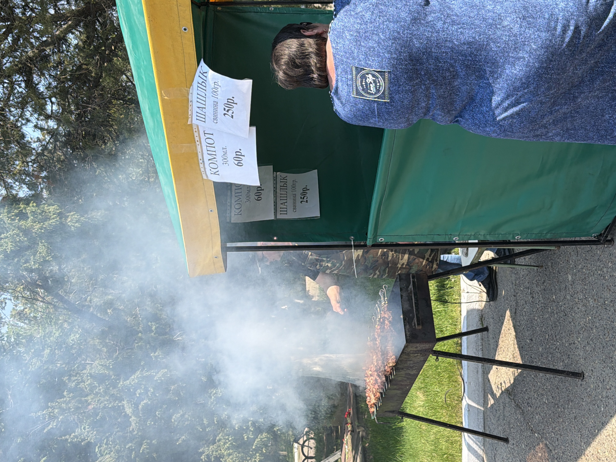 The image shows a street food stall in Kamyshin, Russia. A man wearing a blue shirt with a patch is standing at the stall. The stall has a yellow and green awning and two signs advertising "Компот" (compote) for 60 rubles and "Шашлык" (shashlik) for 250 rubles. Smoke is rising from the grill where the shashlik is being cooked by a person wearing camouflage clothing. The grill is made of black metal and has several skewers of meat cooking on it. The stall is located on a paved area next to a grassy area with trees.