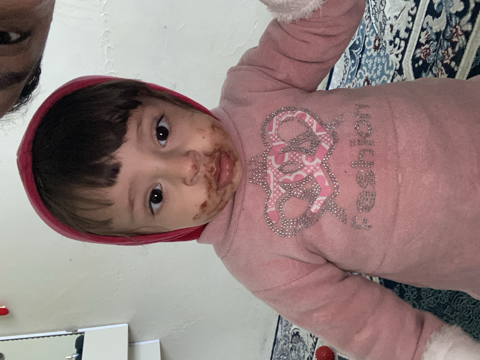 The image depicts an indoor scene within what appears to be a private residence in Kandahar, Afghanistan. The precise location within the city is not discernible from the visual evidence.
A young child, approximately a toddler, with dark hair and bangs, is positioned centrally in the frame, looking directly at the camera. The child wears a red hooded garment and a pink long-sleeved top featuring a rhinestone crown design above the word "Fashion." A dark brown, viscous substance is present around the child's mouth, chin, and lower cheeks.
In the upper right corner, the top of an adult's head, including an eye, is partially visible, indicating close proximity to the child, likely as the photographer.
The background consists of a plain, light-colored wall, possibly off-white, with some minor surface imperfections. To the left, a portion of a white wall-mounted shelf or cabinet is visible, which includes a mirror reflecting an indistinct area. The floor is covered by a patterned rug with blue, white, and darker abstract or floral designs. Lighting is consistent with an indoor setting, producing soft, even illumination. No specific actions beyond the child's direct gaze and the adult's implied presence are overtly depicted.