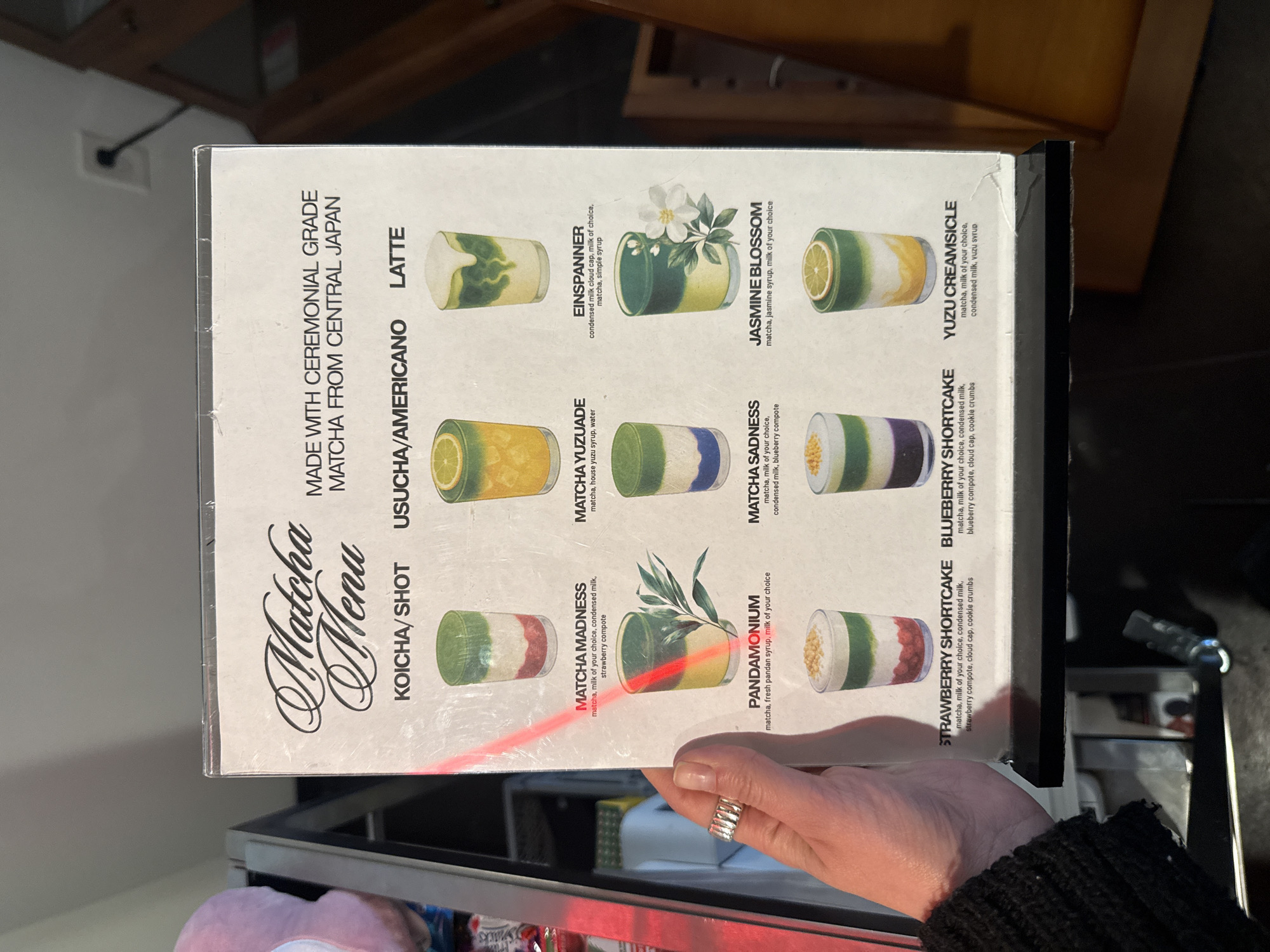 Here is a detailed description of the image:
**Scene:**
The image shows a close-up view of a menu, predominantly white with black text and colorful drink illustrations. The menu is a rigid board framed in black, and a portion of a person's hand holding the edge of the menu is visible. The hand has pale skin and is wearing a silver ring. The person is wearing a dark knit sweater. The backdrop includes shelves with items that are not clearly identifiable, some in packaging with red and pink colors.
**Objects & Details:**
* **Menu:** The menu presents a "Matcha Menu" with various matcha-based drinks, including "Matcha Madness," "Matcha Yuzuade," "Einspanner," "Pandomonium," "Matcha Sadness," "Strawberry Shortcake," "Blueberry Shortcake," "Jasmine Blossom," and "Yuzu Creamsicle." Each drink is briefly described with its ingredients, such as matcha, condensed milk, fruits, and syrups.
* **Text:** The menu specifies that the matcha used is "MADE WITH CEREMONIAL GRADE MATCHA FROM CENTRAL JAPAN." The menu is segmented into categories "KOICHA/SHOT," "USUCHA/AMERICANO," and "LATTE."
* **Background:** The background includes partial views of a shelf or counter area with miscellaneous items.
**Location:**
Without additional information, it's not possible to definitively pinpoint the exact location within Dallas, USA. The menu suggests a cafe or eatery specializing in matcha-based beverages.