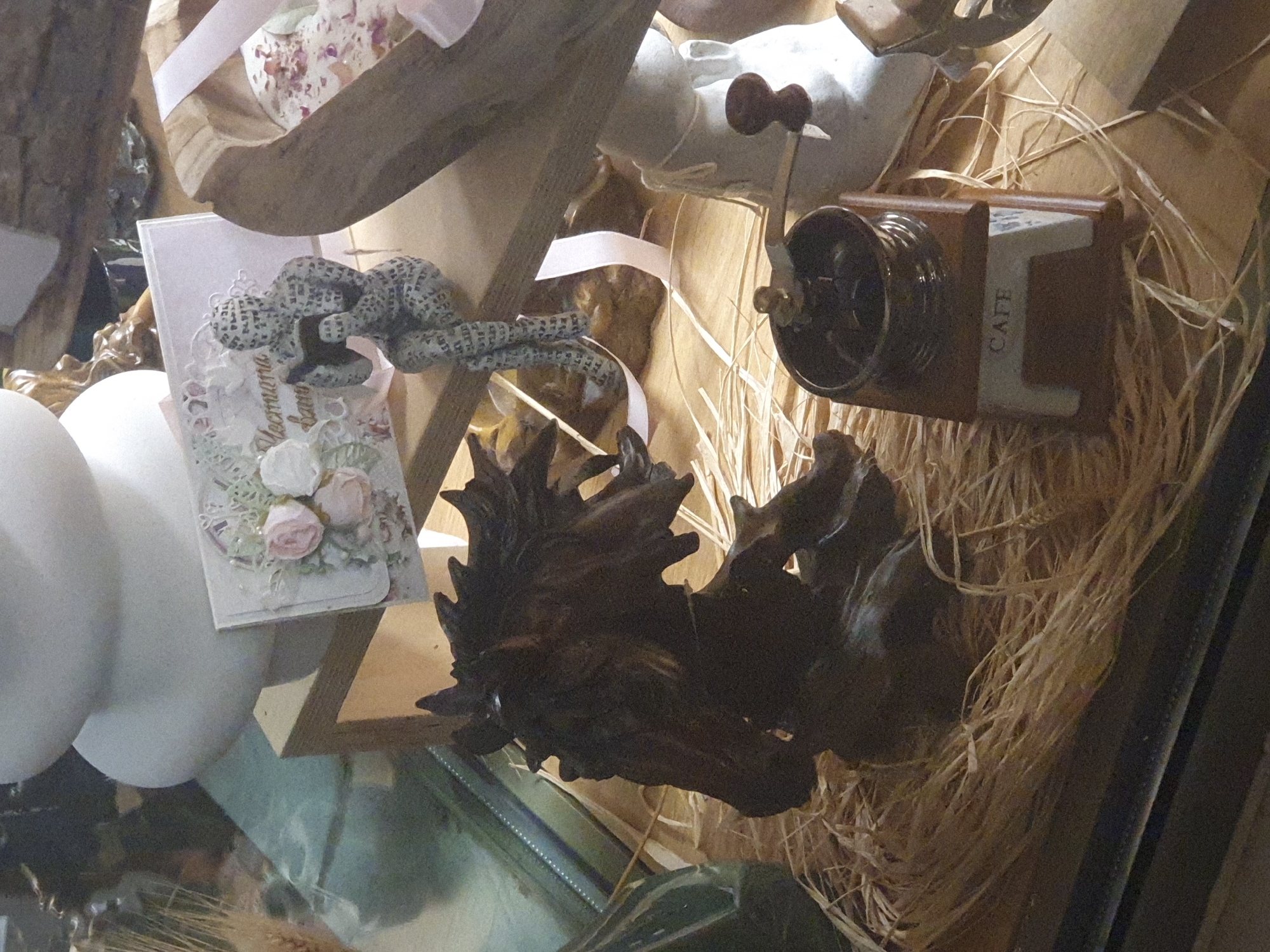 The image shows a collection of decorative items likely arranged inside a shop or gallery. The scene includes a wooden coffee grinder labeled "CAFE" resting on straw, a piece of gnarled wood, a greeting card with fabric roses, and other unidentifiable objects. The location is potentially in Plovdiv, Bulgaria, based on the prompt instructions, but the image doesn't offer any visual confirmation. There are no people visible.