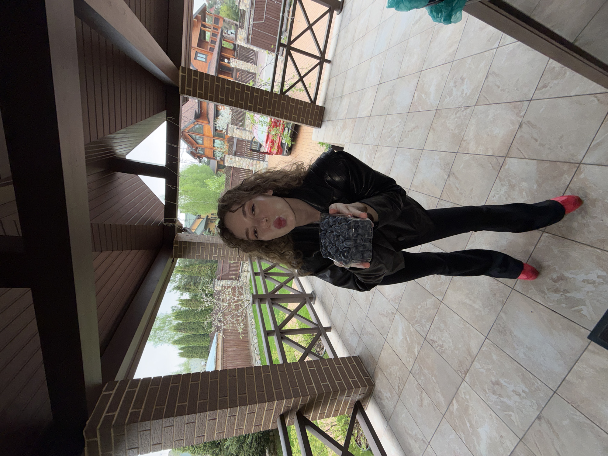 A woman with long, curly brown hair, wearing a black leather jacket and black pants, is holding a clear container filled with dark blueberries in front of her. She is standing on a tiled porch or patio. The tiles are light beige. She is wearing red shoes. Behind her, there is a wooden railing and posts constructed of brick. Beyond the railing is a residential area, with houses and trees visible. The location is Новоглаголево, Russia, as inferred from image tags. Overhead is a brown wooden structure, suggesting a covered porch. The woman is looking at the camera, making a pouting facial expression.