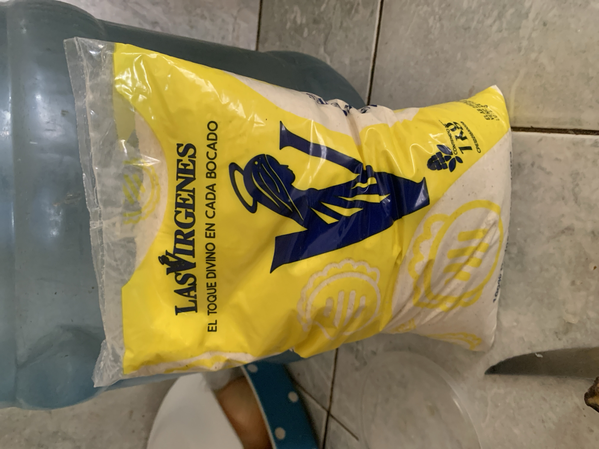 A vertically oriented, close-up indoor photograph displays a yellow and white plastic bag, likely containing flour or sugar, resting on its side. The bag is branded "LAS VIRGENES" in dark blue capital letters, with the slogan "EL TOQUE DIVINO EN CADA BOCADO" (The divine touch in every bite) printed below. A large blue graphic depicts the silhouette of a figure, possibly a woman with flowing hair, holding what appears to be a trumpet or horn, from which rays emanate.
Towards the top right of the bag, the text "CONTENIDO 1Kg" (Content 1Kg) is visible, accompanied by a small graphic of a berry or fruit cluster. Smaller, faint text nearby includes "ELAB: 26/02/2021" (manufacture date) and possibly "VENC: 26/08/2021" (expiration date). Through the translucent plastic, the bag's contents appear as a white, granular substance. A large, translucent blue plastic jug or bottle is partially visible on the left side of the frame. The background consists of light-colored tiled surfaces with dark grout lines, suggesting a utilitarian interior setting, possibly a kitchen or pantry, consistent with a household in Santa Ana, Venezuela. No specific activity is taking place; the items are stationary.