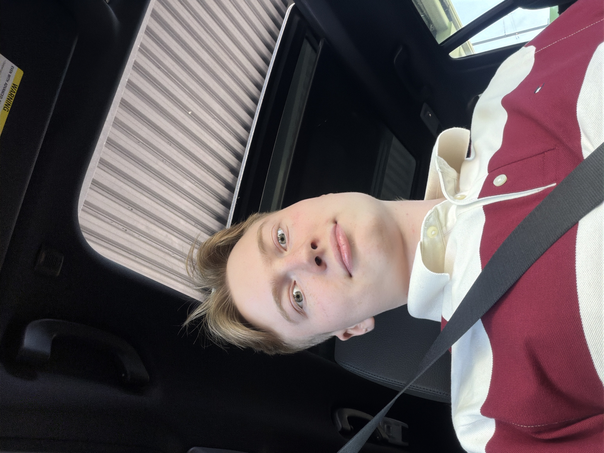 A young man with light brown hair and light skin is sitting in the passenger seat of a car, looking up at the camera. He is wearing a maroon and white striped polo shirt and a black seatbelt. The car has a sunroof with the shade open, revealing a ribbed interior. The scene appears to be outdoors, with some indication of daylight. The image does not provide any specific details about the location in the city of Kyiv, Ukraine.