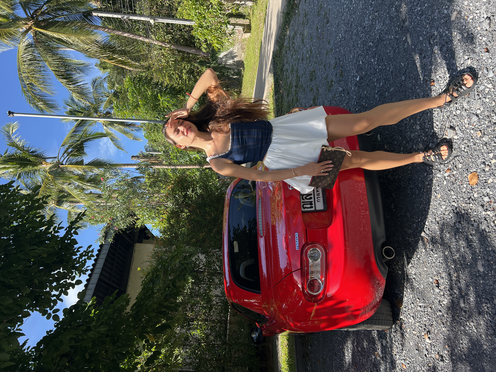 A woman stands next to a red Mazda convertible parked on a gravel surface. She is wearing a denim crop top, a white skirt, and black lace-up sandals. She holds a Louis Vuitton clutch in her left hand and has her right hand on her forehead, shielding her eyes from the sun. Behind her, palm trees and lush green foliage suggest a tropical environment, consistent with the location of Ban Bang Rak, Thailand. A portion of a building with a dark-tiled roof is visible to the left, partially obscured by trees. The sky is bright blue with scattered clouds.