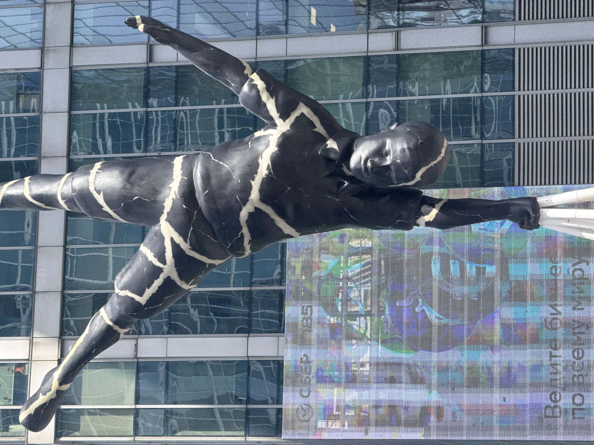 A large, black sculpture of a human figure in a handstand pose with white crack-like markings is positioned in front of a modern glass-faced skyscraper in Moscow, Russia. The sculpture's body is contorted as it balances on one hand, with legs extended upwards. Behind the sculpture, a large digital billboard displays colorful, abstract patterns and text, including Cyrillic lettering that appears to read "Велите бизнес по всему миру" (Manage business around the world). The skyscraper's reflective glass facade shows distorted reflections of the surrounding urban environment. The sculpture is supported by a white tripod structure at its base.