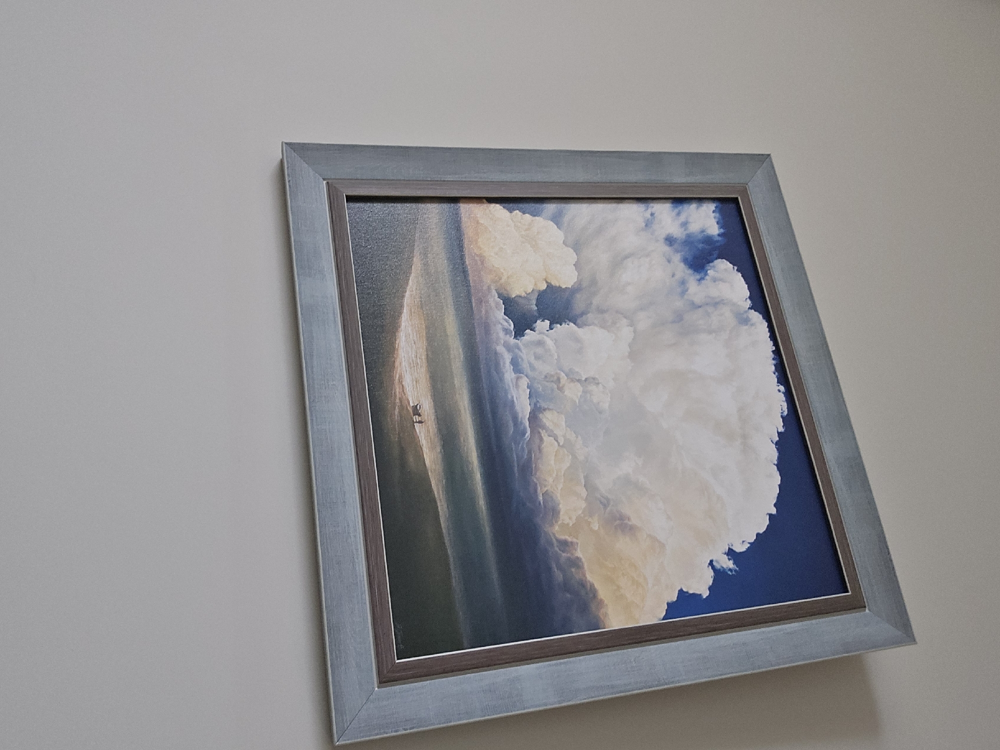 A framed painting or print hangs diagonally on a plain, light-colored wall, likely off-white or light gray. The artwork depicts a dynamic natural scene, possibly a seascape or landscape at sunrise or sunset.
The painting features a vast sky dominated by large, billowing white and subtly colored clouds that transition from bright white to hints of orange, pink, and yellow, suggesting atmospheric light conditions. The upper right portion of the sky is a deep, clear blue, contrasting with the lighter cloud formations. Below the sky, a wide expanse of water or land stretches towards the horizon, reflecting the soft light. A tiny, distant object, which appears to be a sailboat or a small structure, is visible on or just above the horizon line.
The artwork is encased in a double frame. The outer frame has a distressed or washed light blue-gray finish, giving it a somewhat rustic or aged appearance. Inside this, there is a narrower, darker brown or dark gray inner border directly surrounding the image, serving as a mat or part of the frame's design. The photo is taken from a slightly low and angled perspective, causing the painting to appear tilted and receding towards the upper right. No text is visible on the painting or the frame.