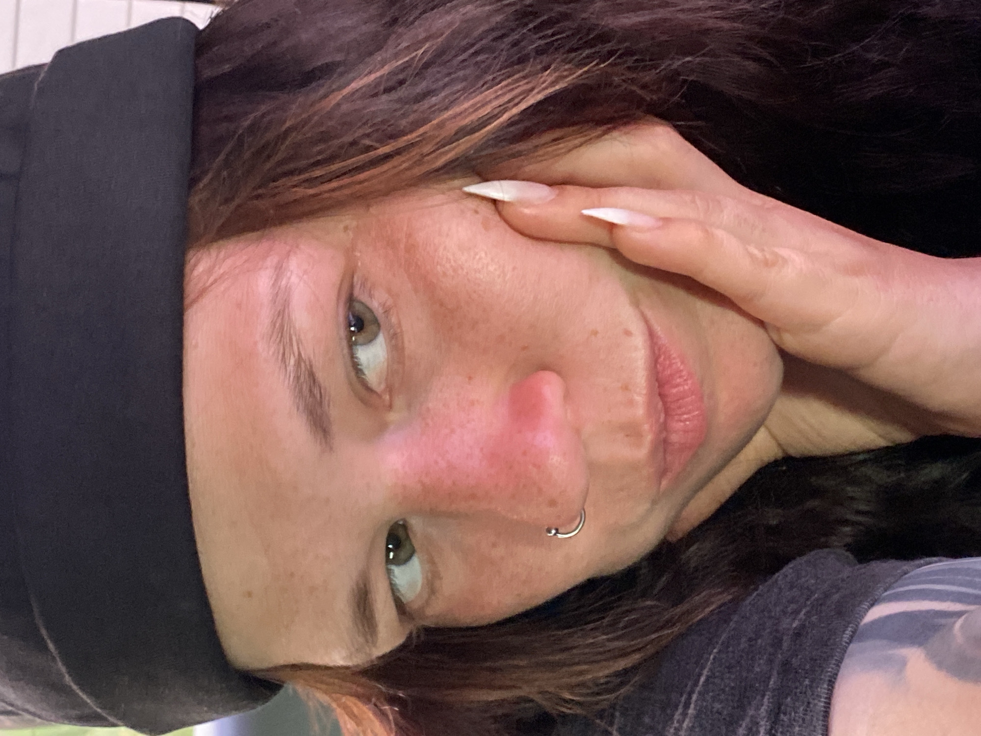 The image is a close-up, self-taken portrait of a person wearing a black beanie and a dark grey t-shirt. The individual has fair skin with freckles, prominent reddish flushing on their cheeks and nose, and long, dark brown hair. They have a silver hoop nose ring. The person is resting their right hand on their cheek, with long, white acrylic nails visible. Their gaze is directed slightly to the left. The background is dark and out of focus. There is no identifiable urban environment present in the image that would allow for a specific location in Верхнеозёрное, Russia to be determined.