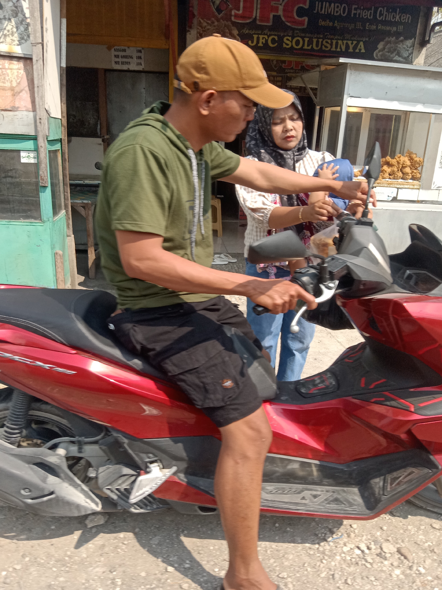 The image shows a man and a woman on a red scooter in front of a food stall in Sugihwaras, Indonesia. The man, wearing a green hoodie and a brown cap, is operating the scooter. The woman, in a hijab and a patterned top, is holding a baby. The food stall appears to be a "JFC SOLUSINYA" which sells "JUMBO Fried Chicken." The stall has a menu board with prices for "NASGOR 10K," "MIE GORENG 10K," and "MIE REBUS 10K." The background shows a building with a yellow facade and windows. The ground is covered in gravel and dirt.
