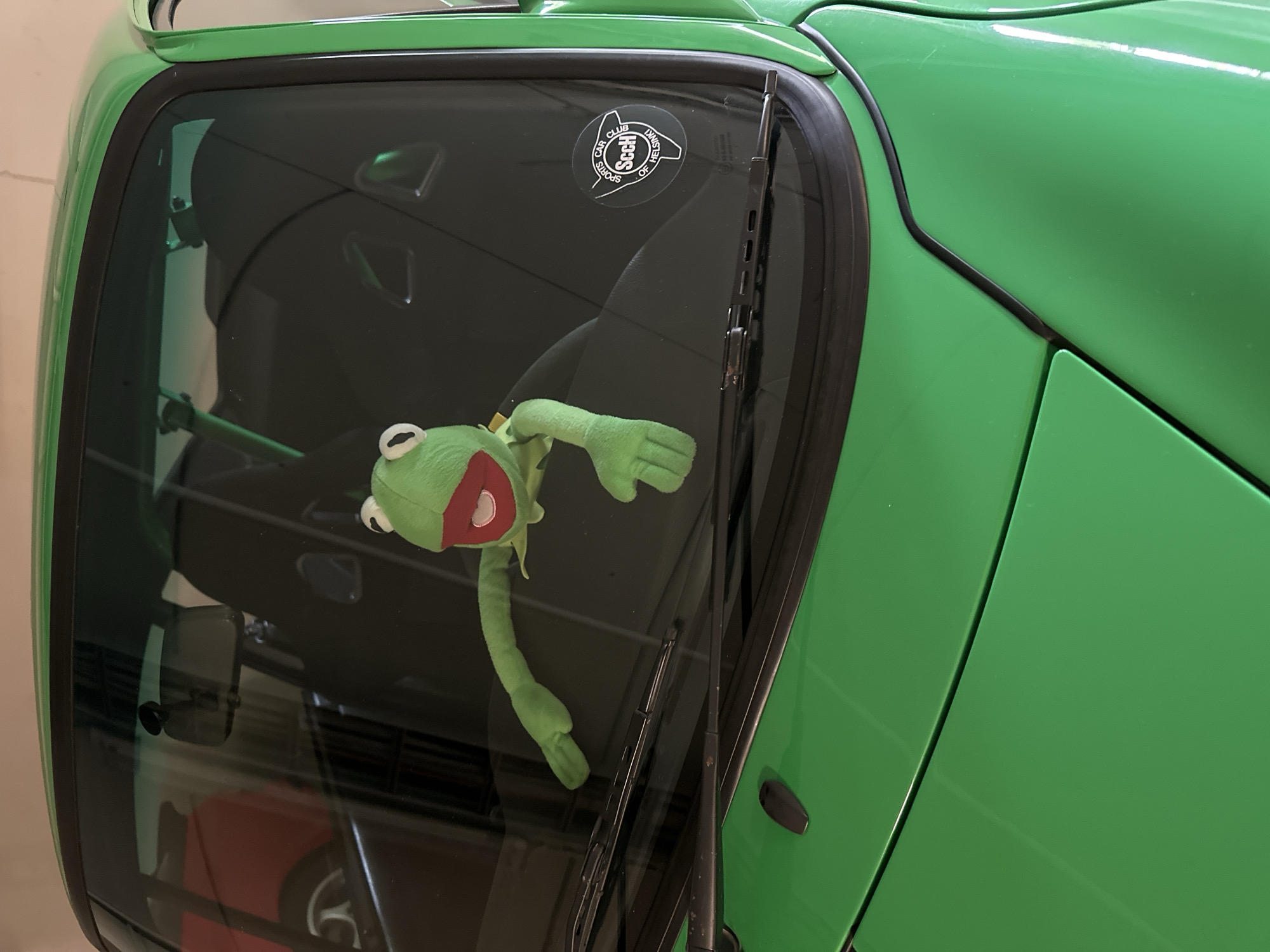 A Kermit the Frog plush toy is positioned on the dashboard of a bright green car, its arms outstretched. The car appears to be a sports car, possibly a Porsche, with black racing seats visible through the windshield. A sticker for the "Sports Car Club of Helsinki" is affixed to the lower right corner of the windshield. The scene is illuminated by artificial light, suggesting it is indoors, likely in a garage. The background is blurred, but appears to show other vehicles and storage shelving, consistent with a private garage setting in Espoo, Finland.