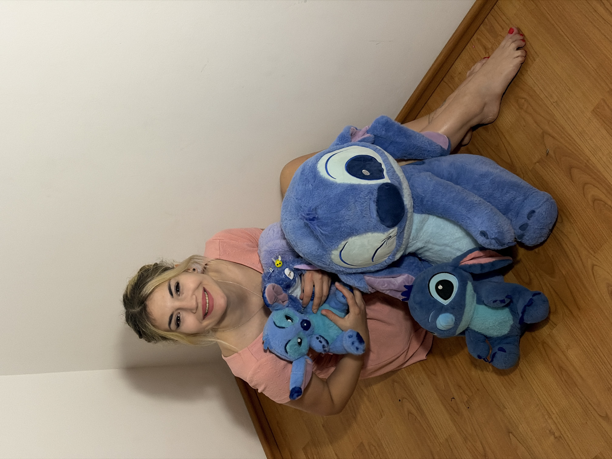 This image, taken indoors in Budva, Montenegro, features a young woman surrounded by several plush toys. The scene is set against a light, off-white wall and a light brown wooden floor, suggesting a cozy room corner.
The central figure is a woman with blonde hair, pulled back from her face, and a fair complexion. She is wearing a light peach or pink short-sleeved top and is depicted lying or seated on the wooden floor, leaning against the wall with her legs extended. Her toes are painted with bright red nail polish. She is smiling widely, looking directly at the camera, conveying a cheerful and content emotion.
She is accompanied by at least three, distinct blue plush toys of the character Stitch from Disney's "Lilo & Stitch." A large Stitch plush rests beside her, and she holds a medium-sized one in her arms. Another smaller Stitch plush is visible on the floor near her. The toys are varied in size but all display Stitch's signature blue color and alien features. The lighting appears to be soft, even indoor illumination. There is no visible text in the image.