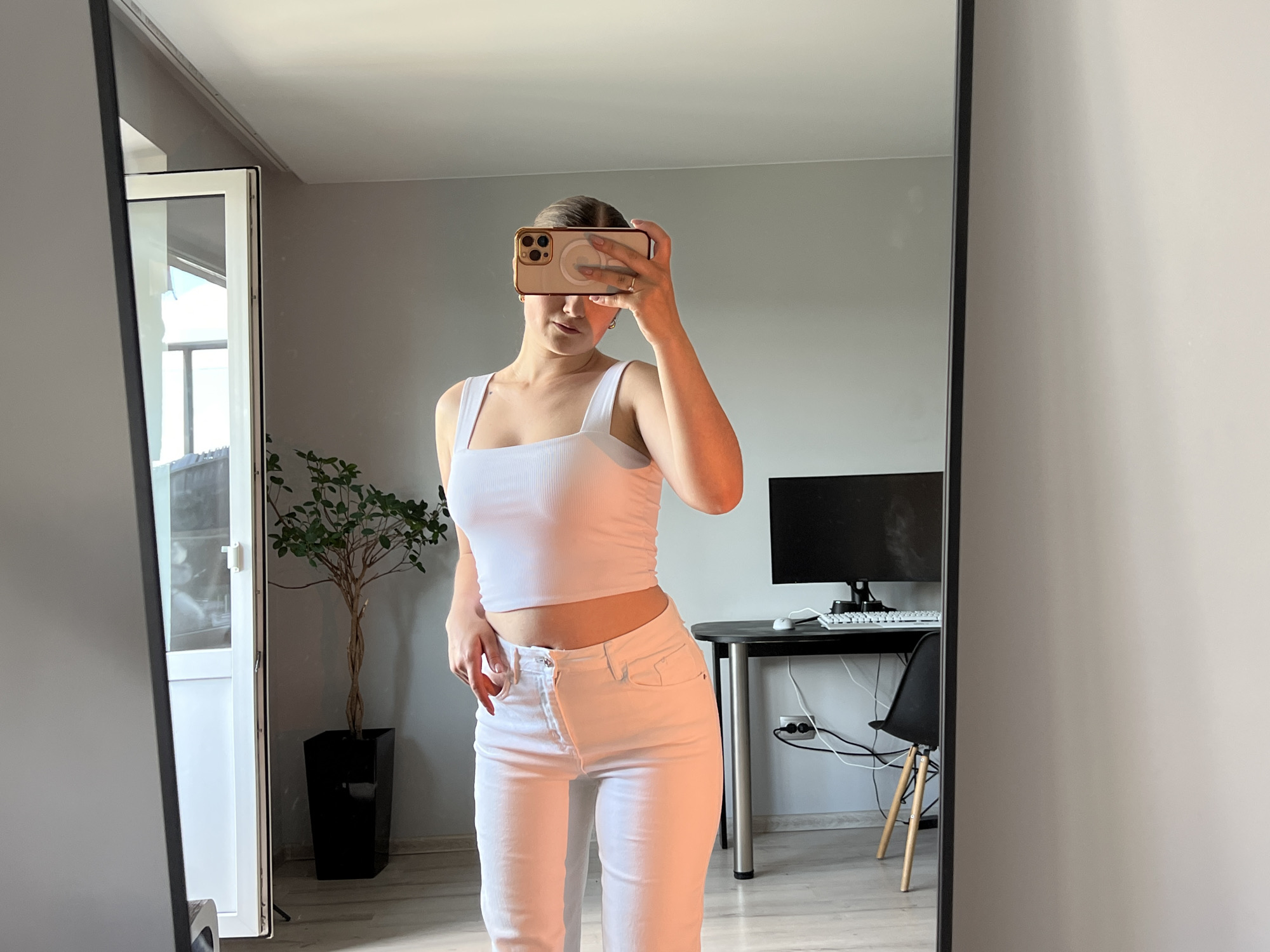 A young woman is taking a mirror selfie in a room. She is wearing a white cropped tank top and white jeans. She is holding a smartphone in front of her face, obscuring it. The room has light gray walls and light wood-grain laminate flooring. To the left, a glass door leads outside to a balcony or patio. A potted plant with green leaves sits next to the door. To the right, a black desk with a computer monitor, keyboard, and mouse is visible, along with a black chair with wooden legs. The mirror itself is large and framed in black. The scene appears to be an indoor residential setting.