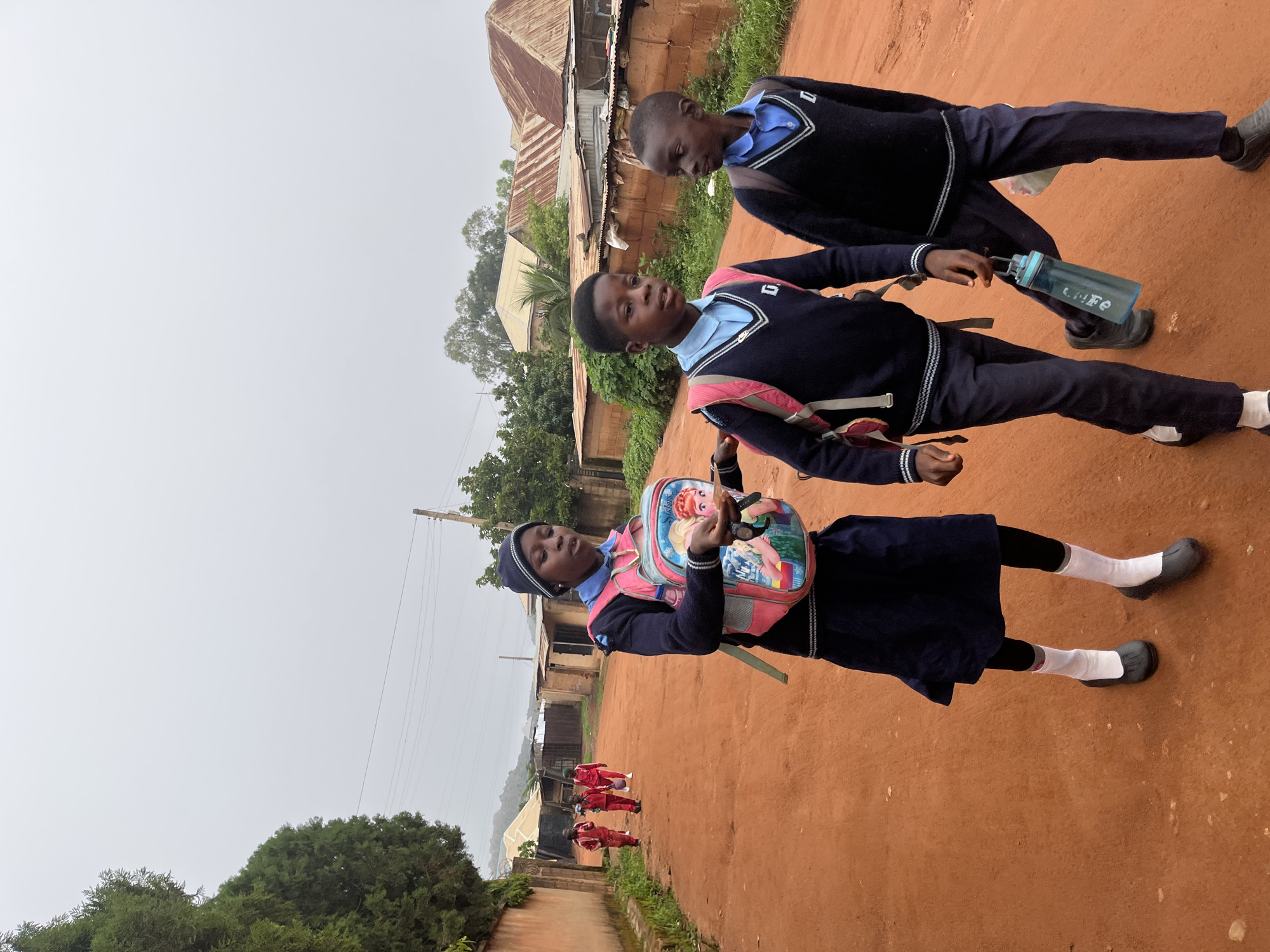 In Jos, Nigeria, on an overcast day, three schoolchildren walk along a dusty, reddish-brown dirt road. The two children in the foreground are a girl and a boy, both wearing navy blue sweaters over light blue collared shirts, navy blue pants, and dark shoes with white socks. The girl, on the left, carries a pink backpack adorned with cartoon characters and has a dark beanie on her head. She is looking down, holding a white object and a pair of sunglasses. The boy beside her carries a similar navy blue sweater and light blue shirt uniform, with a pink backpack slung over his shoulder. He holds a blue water bottle in his right hand. Further down the road, three other children in red school uniforms walk away from the camera. The road is lined with modest buildings with corrugated metal roofs and brick walls, interspersed with trees and vegetation. A concrete wall runs along the left side of the road, separating it from a cluster of trees. Electrical wires are visible overhead, suggesting the presence of infrastructure. The overall scene depicts a typical school day in a Nigerian community.