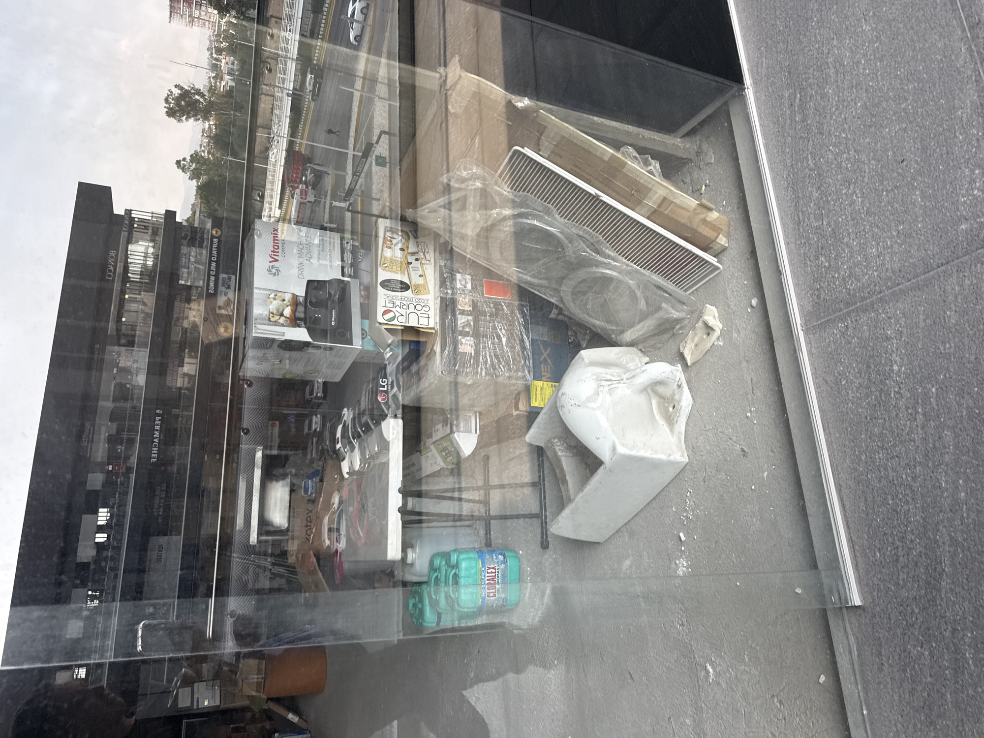 The image shows a window display or storage area with various household items visible, reflected against the outside scenery in San Bernardino Tlaxcalancingo, Mexico. Among the items present are boxes for a Vitamix blender and EURO Gourmet products, a package of Kotex feminine products, an LG product, a package of CLORALEX, and a white ceramic object that looks like a sink or basin. The reflection shows a city street with vehicles and buildings. The area behind the glass also appears to have cardboard boxes and other miscellaneous objects.