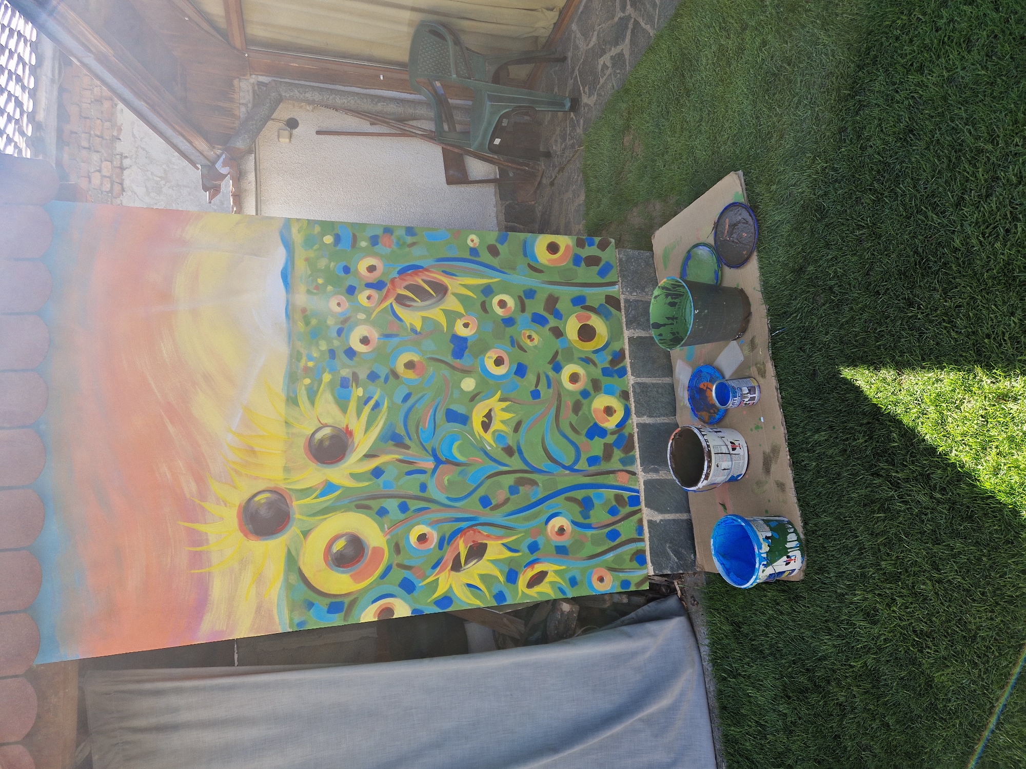 The image shows a painting propped against a structure, possibly a wall or building, in an outdoor setting. The painting appears to be an abstract floral design, featuring sunflowers, with a blend of colors including yellow, green, blue, orange, and brown. The left side of the painting transitions into an abstract sunrise or sunset design.
Next to the painting, on a piece of cardboard on top of grass, are several paint containers of various colors, including blue, green, black, and white. There are also some paint lids.
In the background, a green plastic chair is visible, placed on a stone-paved area, and beyond that is a patch of green grass. Overhead, parts of a building structure with a roof are seen.
The image does not contain information about the specific city or people.