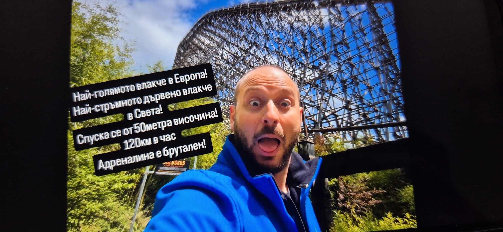 A dynamic medium shot captured from a screen depicts a man posing in front of a massive wooden roller coaster structure. The man, with a bald head and a short beard, wears a blue jacket and makes a comically exaggerated expression of surprise or excitement, with wide eyes and his tongue sticking out. He is positioned slightly off-center to the right, looking directly at the viewer.
Behind him, the intricate lattice work of a large wooden roller coaster dominates the upper and left portions of the background, contrasting with the bright blue sky visible through its framework. Green foliage and trees are visible beneath and around the roller coaster structure.
Several black text bubbles with white Bulgarian text are overlaid on the image, providing details about the roller coaster:
* "Най-голямото влакче в Европа!" (The biggest roller coaster in Europe!)
* "Най-стръмното дървено влакче в Света!" (The steepest wooden roller coaster in the World!)
* "Спуска се от 50метра височина!" (It descends from a 50-meter height!)
* "120км в час" (120 km per hour)
* "Адреналина е брутален!" (The adrenaline is brutal!)
A smaller, partially visible sign near the bottom left has the text "MULUSSOS" (likely "Colossos").
The image appears to be a photograph of a digital display, indicated by the visible screen borders, slight reflections, and mild pixelation. The overall mood conveyed is one of thrilling adventure and excitement, likely at an amusement park during daylight hours.