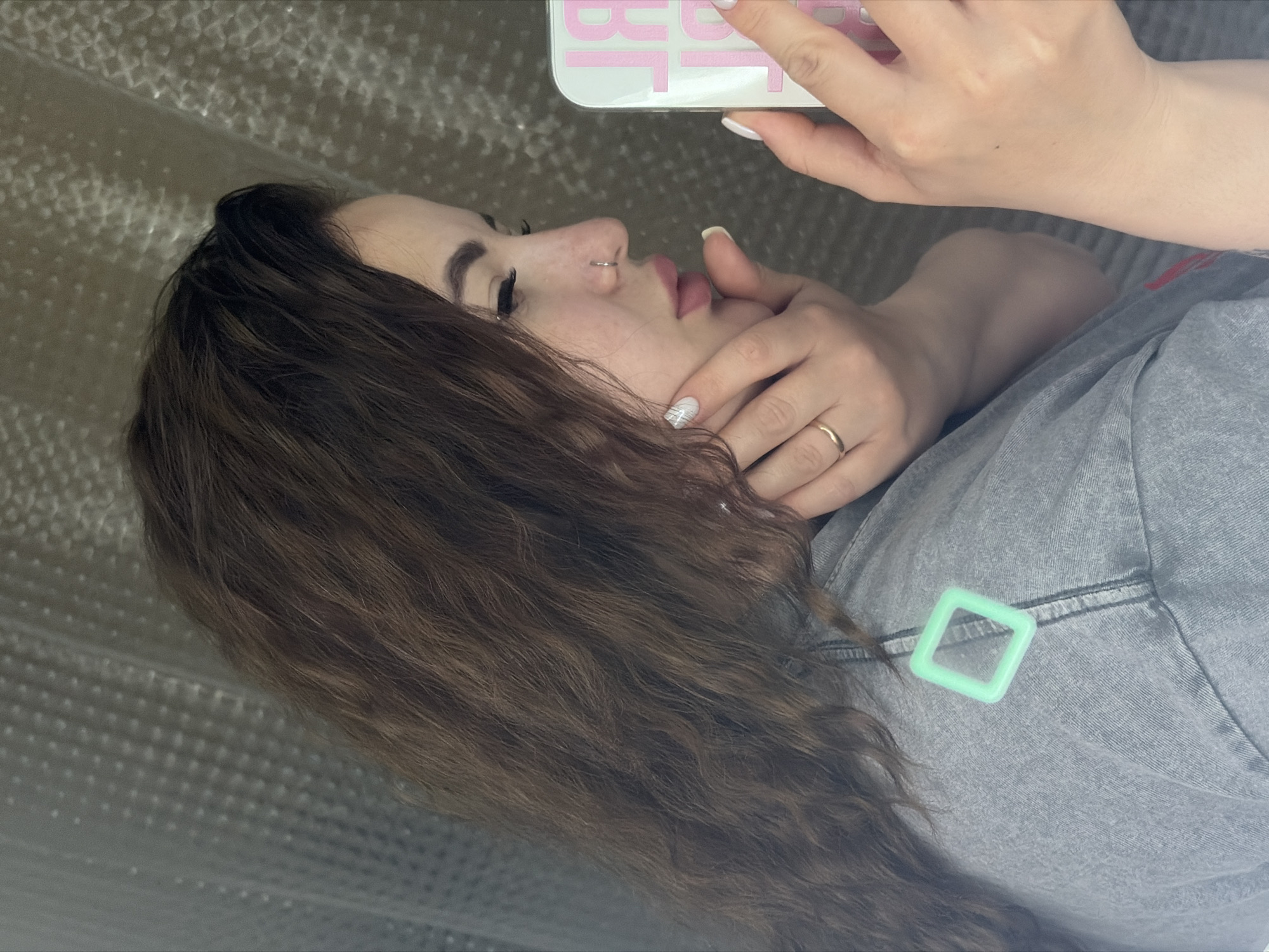 A woman with curly brown hair, a nose ring, and a gold ring on her finger is holding a phone with a white and pink case. She is wearing a gray denim jacket with a square green accessory attached. The background is a textured, light-colored surface. The location is not identifiable from this image.