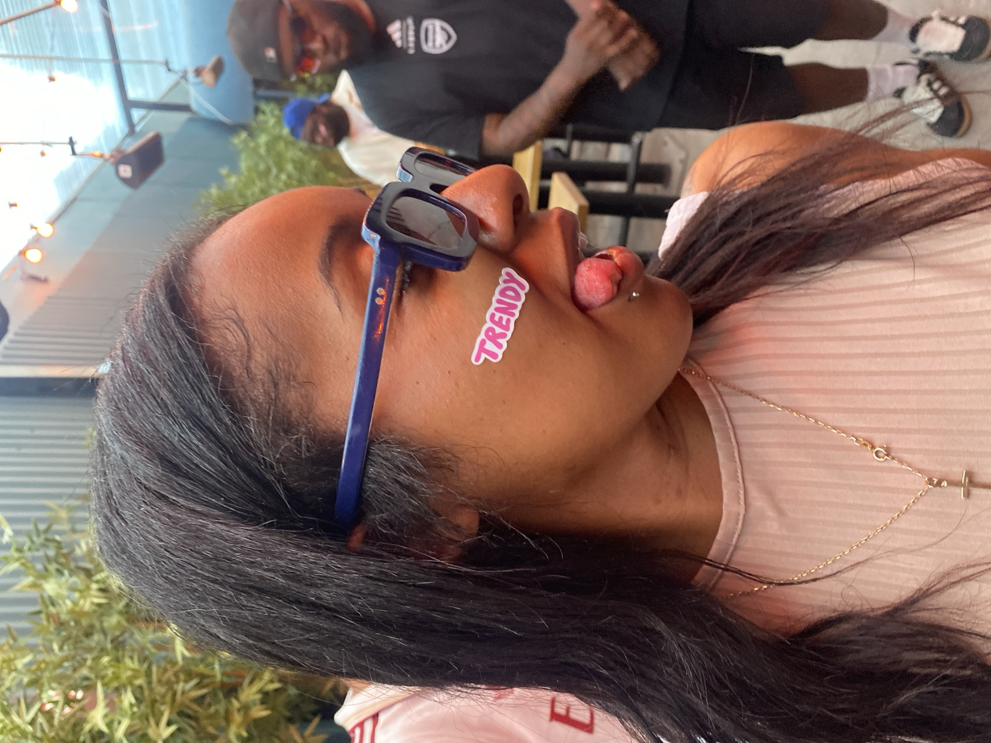 The image shows a dark-haired woman with sunglasses and a "Trendy" sticker on her face, sticking out her tongue. She's wearing a light pink top and a gold necklace with a cross pendant. In the background, two men are visible: one wearing a black shirt and sunglasses, and another wearing a blue cap. The setting appears to be outdoors, with a building's exterior and some greenery visible, but the exact location within London, United Kingdom, cannot be determined from the image.