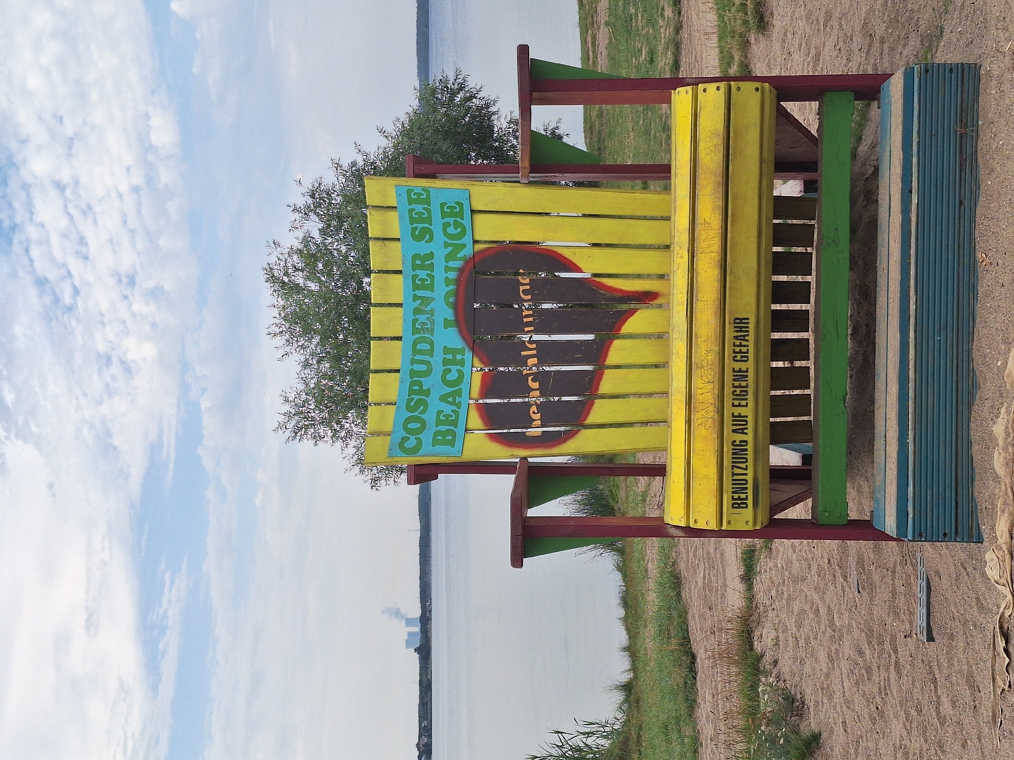 The image captures a humorous, oversized Adirondack-style chair painted yellow, red, and green, positioned on a sandy area near a lake. A teal banner across the top of the chair reads "COSPUDENER SEE BEACH LOUNGE." Below the banner, a dark heart shape is spray-painted, with the word "beach" inscribed within it. Another black banner at the bottom of the chair states, "BENUTZUNG AUF EIGENE GEFAHR," which translates to "USE AT YOUR OWN RISK." In the background, the calm, grey-blue water of a lake stretches towards a hazy horizon. On the far left, distant industrial cooling towers with plumes of smoke are visible against a cloudy sky. The foreground shows dry grass and sand. The chair's exaggerated size and playful colors suggest it's a decorative or novelty item intended for public amusement. The location is the Cospudener See, a lake in Leipzig, Germany, known for its recreational activities.