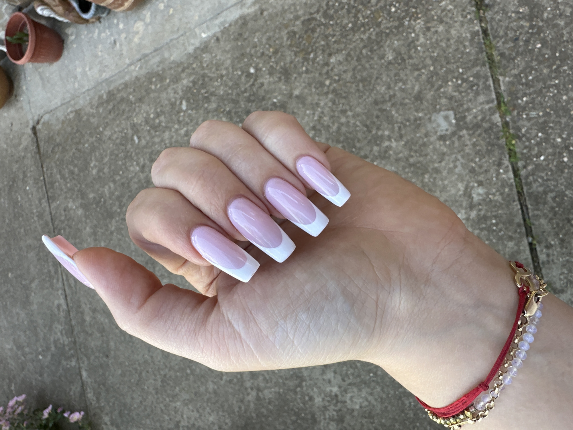 A female hand is displayed against a gray concrete background. The hand's nails are manicured in a French style with a light pink base and white tips. The hand wears two bracelets on the wrist, one gold chain and one red string with pink beads. The background is concrete with a few plants visible. The image may be taken in a yard or outdoor area. It is not possible to verify the location as Lehchevo, Bulgaria.