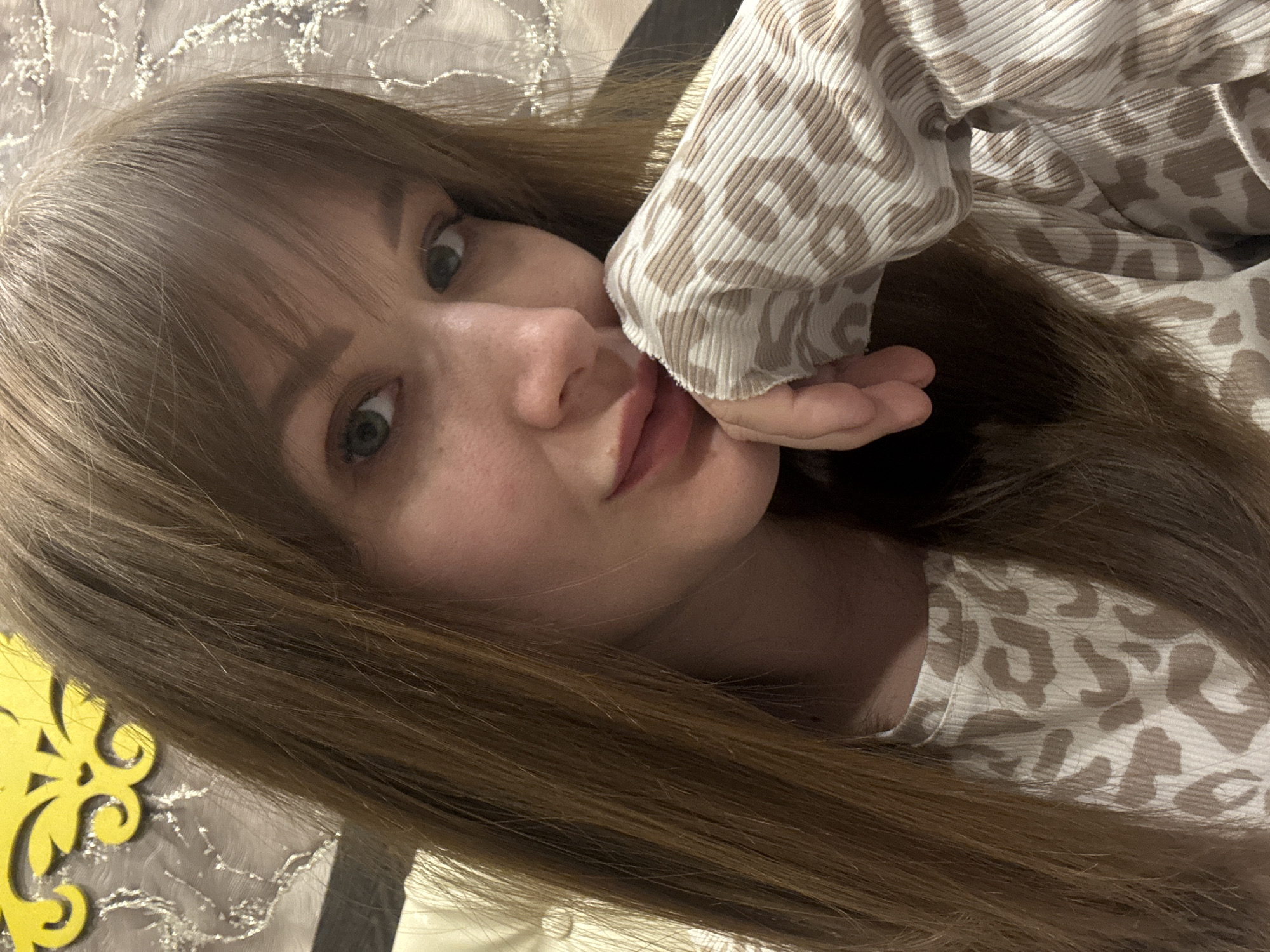 A woman with long, light brown hair is posing for a selfie. She has bangs and green eyes. She is wearing a light-colored top with a brown animal print. Her left hand is up by her face, her fingers slightly curled. The background appears to be a bed with a patterned sheet. The image does not contain sufficient information to determine the location in the city of Chaikovsky, Russia.