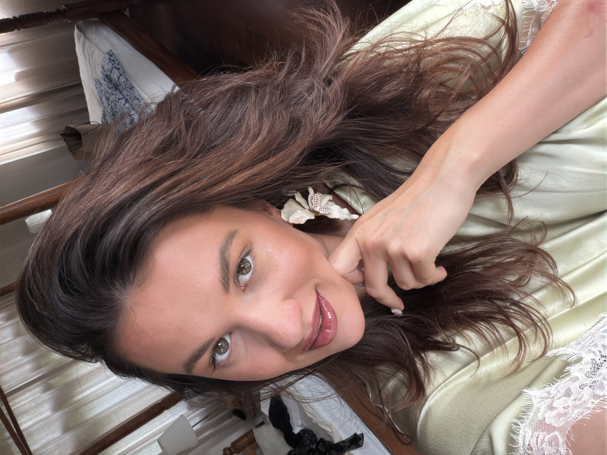 In a bedroom setting, a woman with long, brown, wavy hair is shown in a close-up shot, looking towards the camera. She is wearing a light green satin robe with lace detailing on the shoulder. Her right hand is resting on her chin, and she is wearing a large, white, floral-shaped earring on her left ear. The background reveals a bed with white sheets and a blue patterned blanket, along with a wooden four-poster bed frame and a lamp on a nightstand. Curtains are visible behind the bed. The image is likely taken in North Kuta, Indonesia, given the context provided.