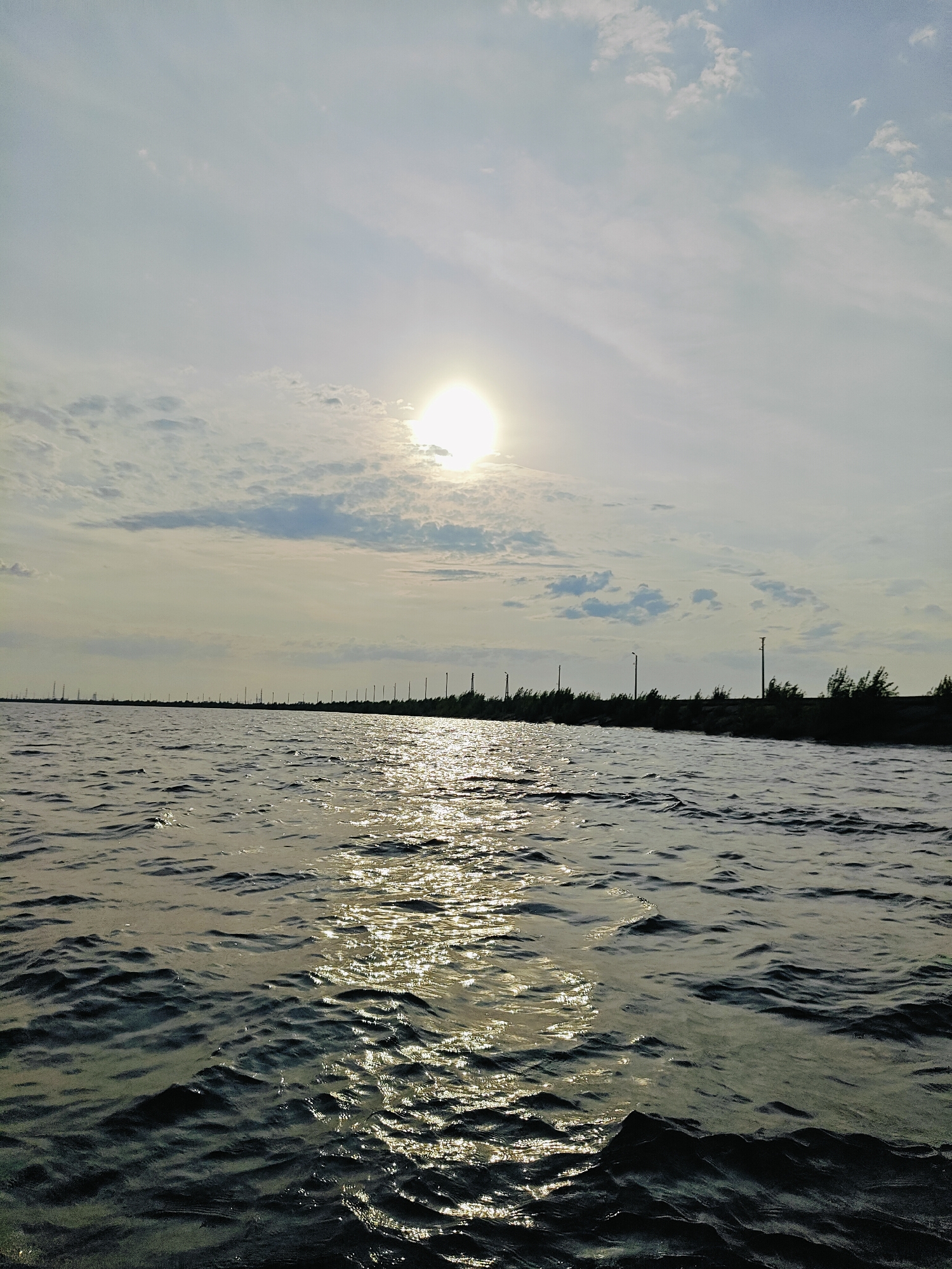 The image shows a body of water, likely the Kama River, with a distant shoreline. The sun is visible in the sky, partially obscured by clouds, casting a bright reflection on the water's surface. The shoreline is lined with trees and what appear to be utility poles or light posts, suggesting a developed area. The water exhibits small waves and ripples, indicating a gentle breeze. The overall scene conveys a sense of calm and natural beauty, characteristic of a riverside landscape in Chaikovsky.