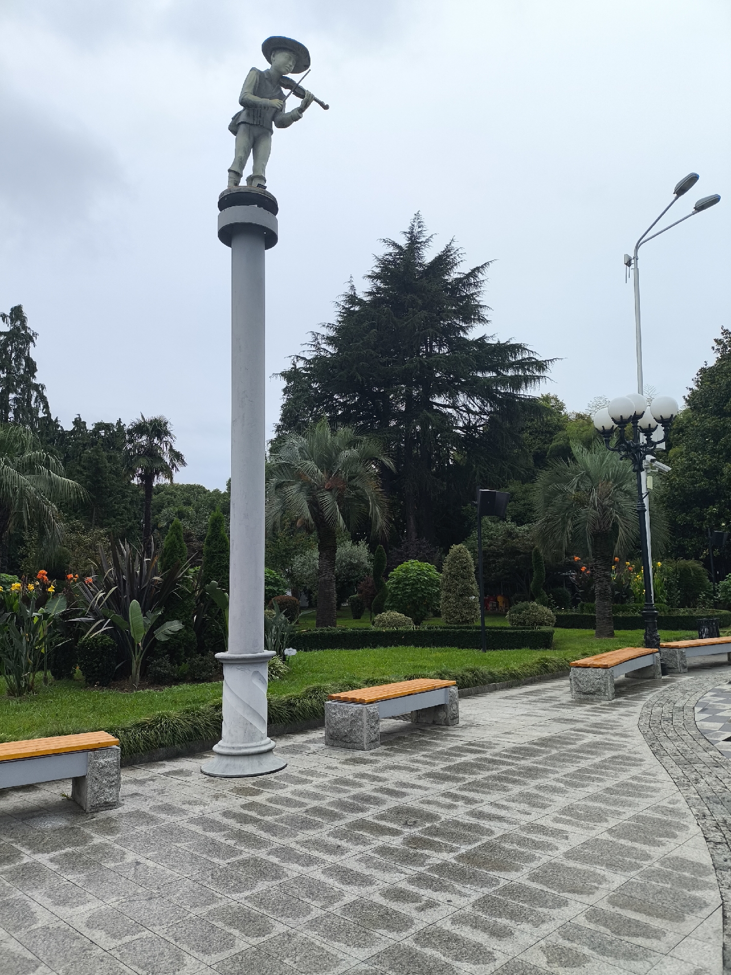 The image shows a paved walkway in Batumi, Georgia, likely in a park or public garden. A statue of a young boy playing the violin stands atop a tall, grey lamppost in the foreground. The walkway is wet, suggesting recent rain. Benches with wooden seats and stone bases are placed along the path. Lush greenery, including palm trees and various shrubs, forms the backdrop, with a large evergreen tree to the left and a more distant street lamp on the right. The sky is overcast.