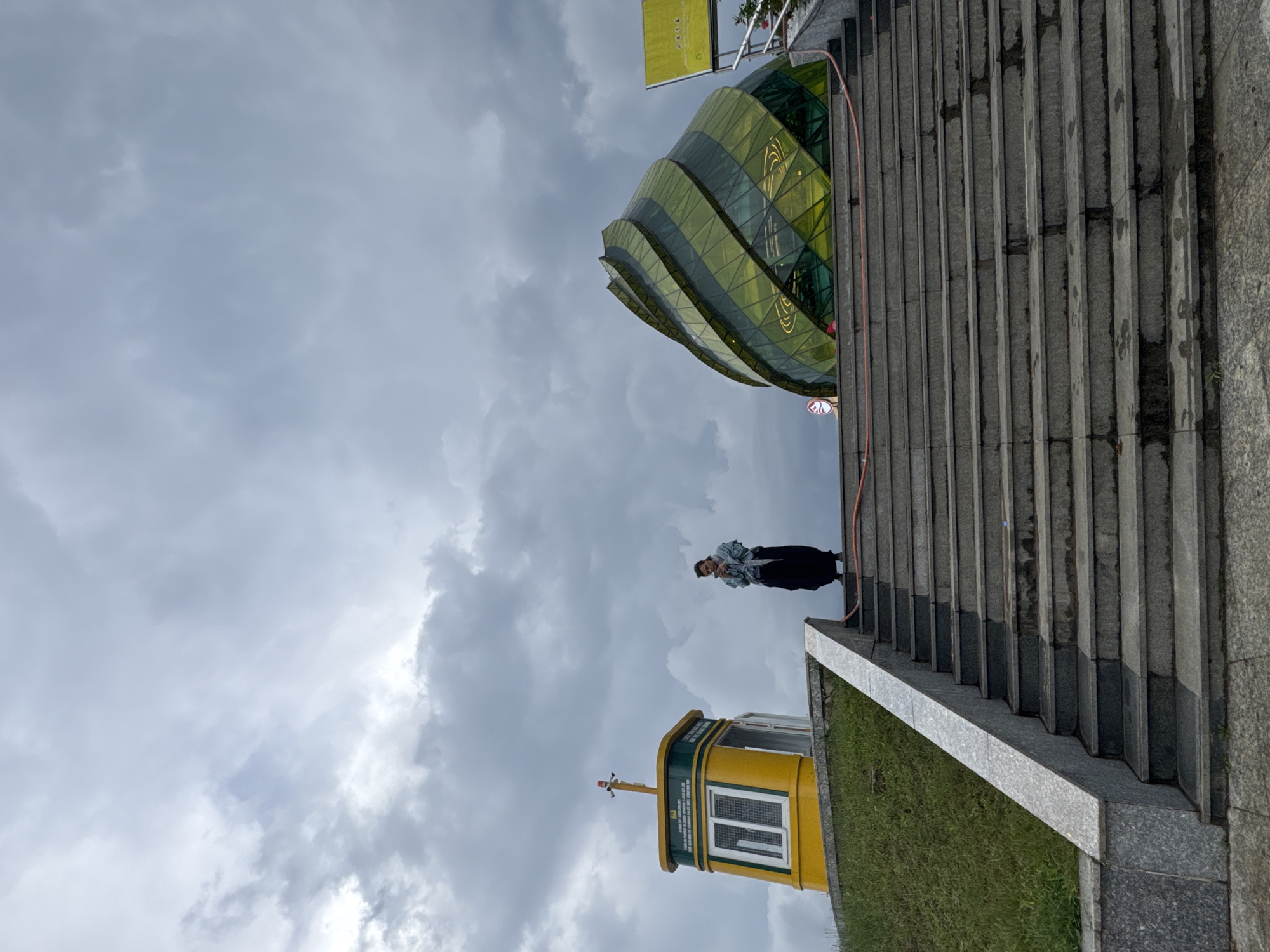 A photograph captures a person standing on a concrete staircase, with a unique structure behind them against an overcast sky. The building is a cylindrical glass structure. Nearby, a yellow tower-like structure is visible, its architecture suggesting a potential cable car station. The location is identified as Da Lat, Vietnam.