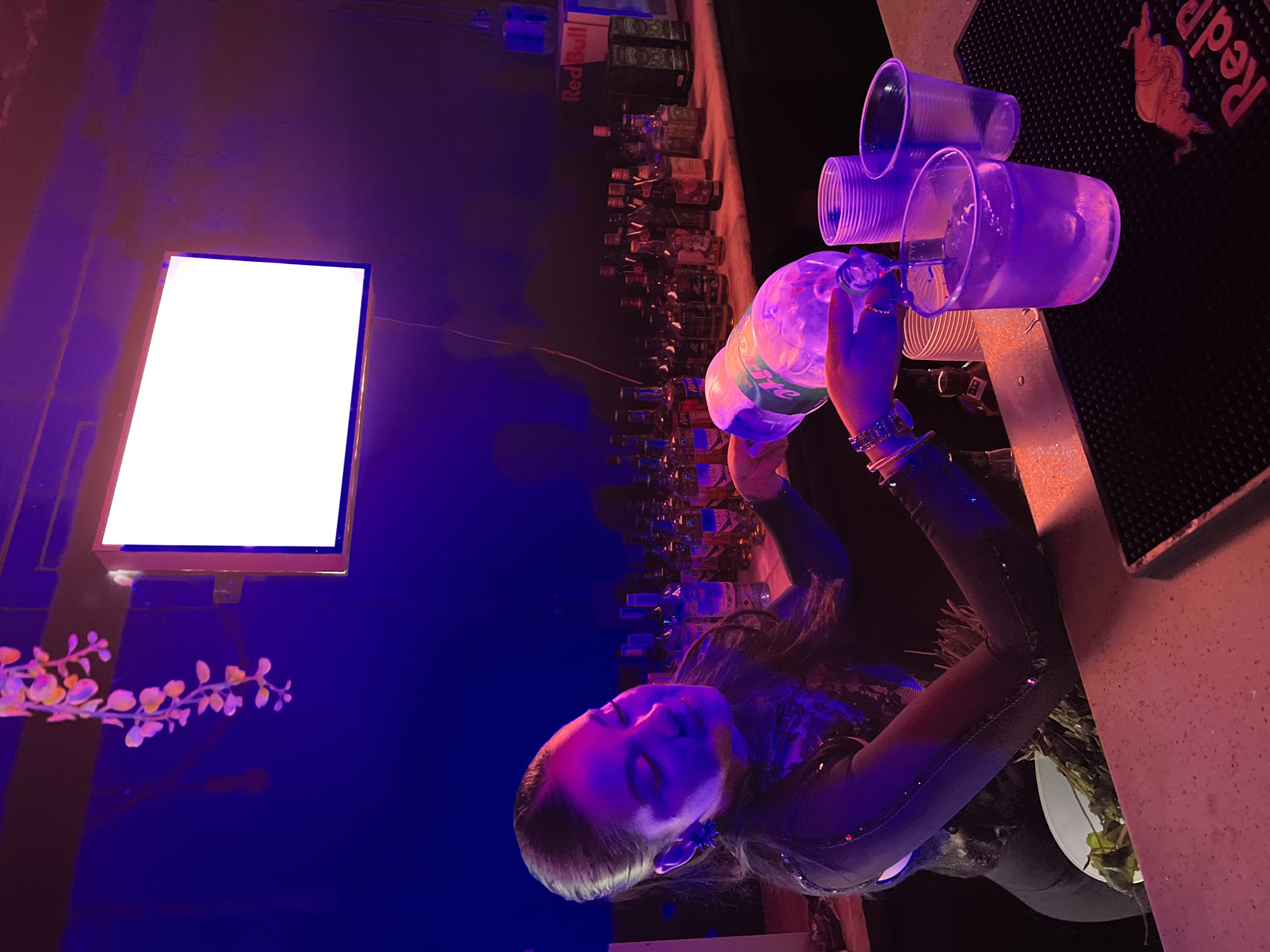 The image shows a woman working at a bar, pouring a clear liquid from a plastic bottle into a stack of clear plastic cups. Behind her, a backlit display screen is visible. Shelves of liquor bottles line the back wall. The bar surface features a Red Bull-branded mat. The image has strong purple and blue lighting, typical of a nightclub setting. The location cannot be identified as Arica, Chile, based on the visual information.