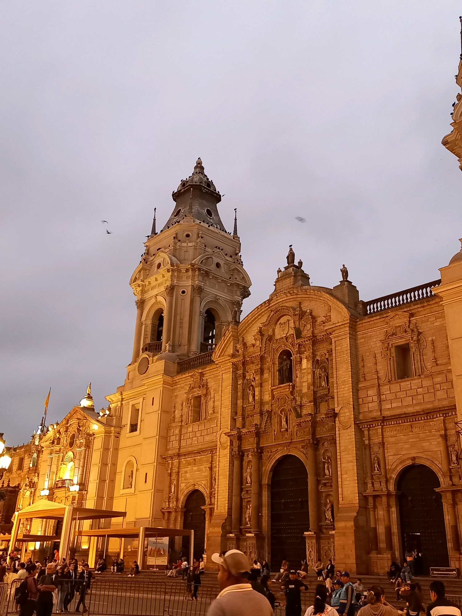 The image depicts the Lima Cathedral, a Roman Catholic cathedral located in the Plaza Mayor in Lima, Peru, at dusk. The grand facade of the cathedral, illuminated by warm artificial lighting, showcases intricate architectural details, including multiple arched doorways and ornate carvings. A prominent bell tower rises to the left of the main structure. The sky above is a muted gray, with a few birds in flight. In the foreground, a crowd of people are gathered, some standing behind a metal barrier, while others are seated on the steps of the cathedral or walking in the plaza. Temporary structures with canopies are also visible in the area in front of the cathedral. The scene suggests a public gathering or event taking place in the historic city center.