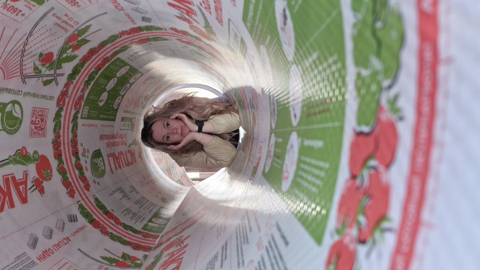 A Caucasian woman with long, wavy blonde hair lies inside a large, cylindrical object adorned with promotional images for greenhouse products. She rests her chin on her hands, appearing relaxed. The interior surface of the cylinder features green and red designs including images of tomatoes, text in Russian advertising greenhouse advantages, and the word "ACTUAL!" The outside environment is faintly visible behind her, implying a bright outdoor setting, likely in Otchor, Russia given the Russian text.