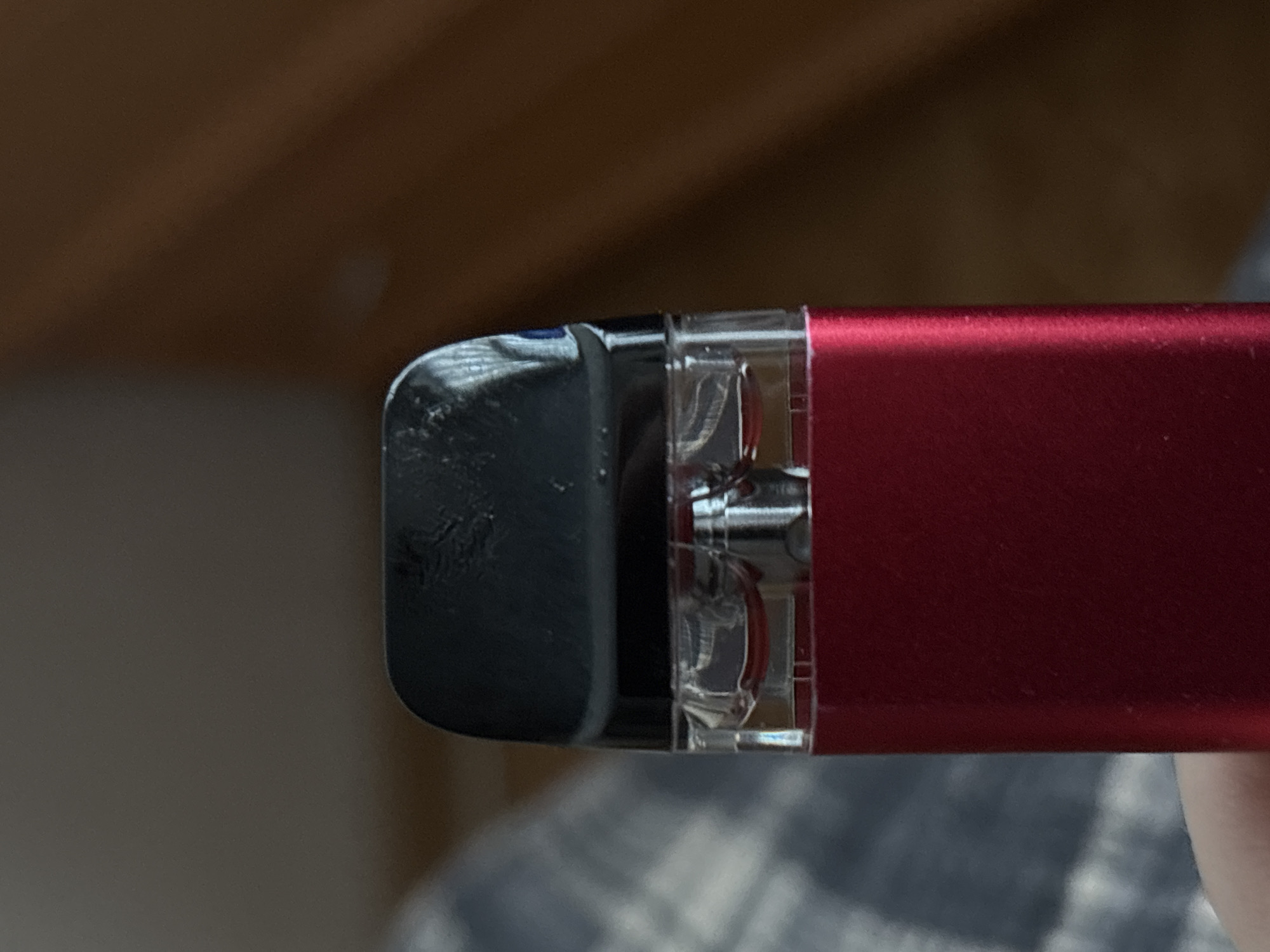 The image is a close-up, eye-level, indoor shot of the top portion of a red vape pen. The vape pen has a black, glossy mouthpiece. Below the mouthpiece, a clear plastic cartridge is visible, containing a small amount of red liquid. The red cartridge is attached to the main body of the vape pen, which is also red and appears to be made of metal. The background is out of focus and consists of muted colors, suggesting an indoor setting with soft lighting. To the left of the vape pen, a blurred pattern of blue and grey fabric is visible, possibly a blanket or clothing. To the right of the vape pen, the background is a solid, dark brown color, hinting at a wall or furniture. No people are visible in the image, and no specific location in Saint Petersburg, Russia, can be identified.