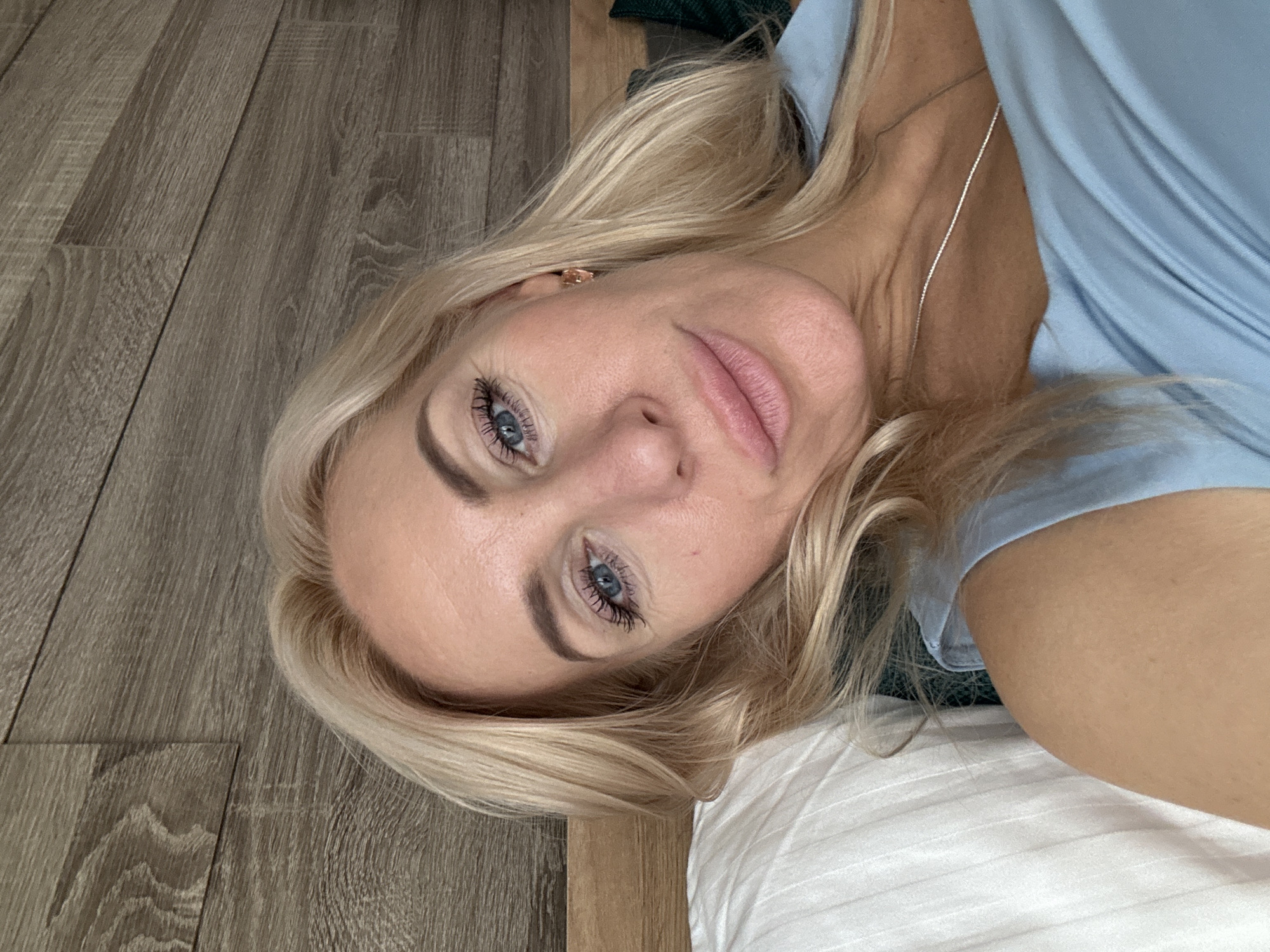The media file is a portrait of a blonde woman lying down. She has blue eyes, full lips, and light skin. She is wearing a light blue top and a silver necklace. Her hair is blonde and falls around her face. The background consists of a wooden floor and a white surface, possibly a bed or pillow. The image is taken from a close-up perspective, focusing on the woman's face and upper body. The lighting suggests it is daytime. There is no visible text in the image.