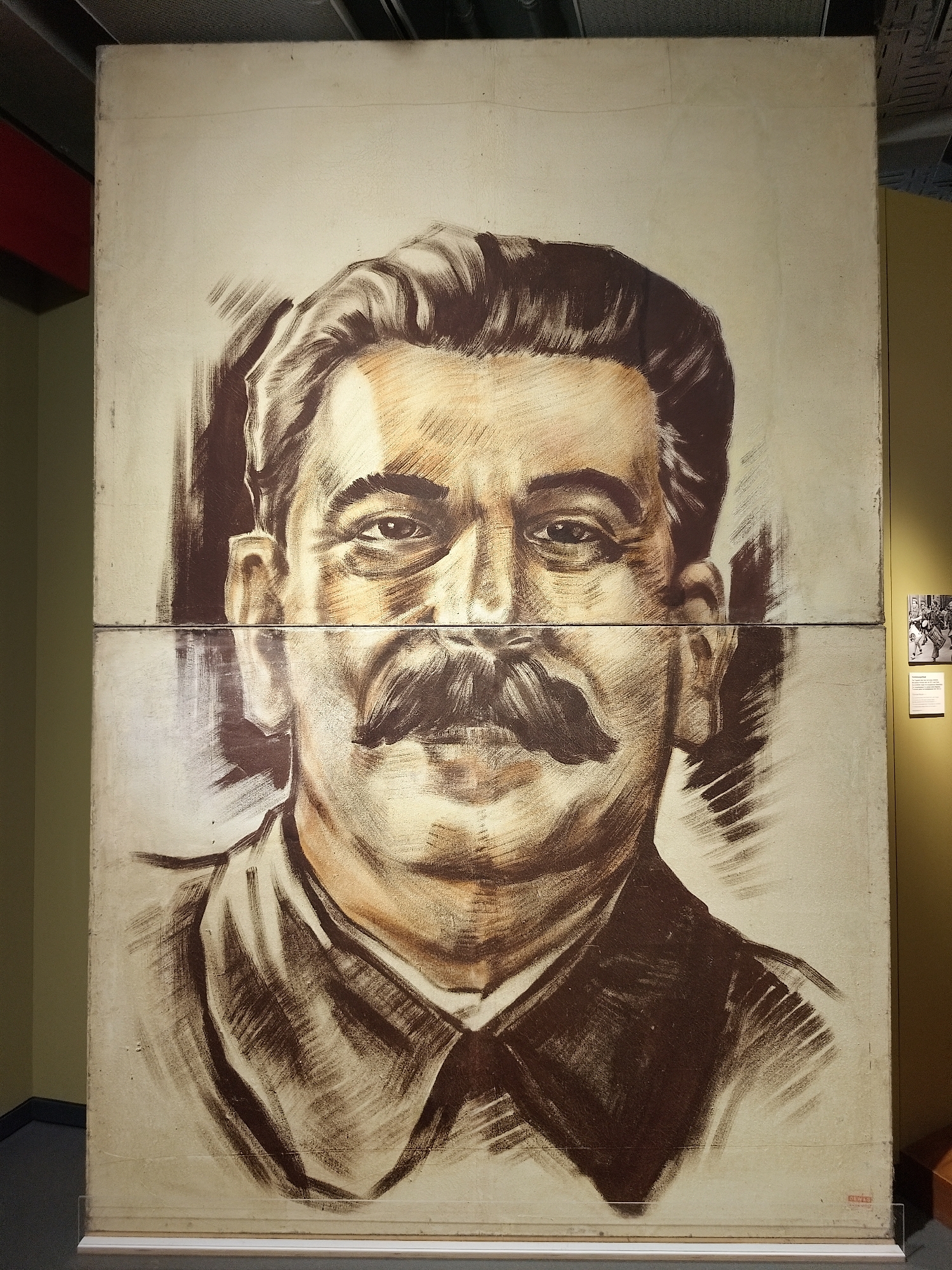 The image is a large, seemingly painted portrait of Joseph Stalin, composed of two horizontal panels. The artwork features a head-and-shoulders depiction of Stalin with a dark mustache and hair, rendered in shades of brown and tan on a beige background. The portrait appears to be displayed in a museum or gallery, as suggested by the lighting, the presence of a small sign and photograph on the adjacent wall, and the overall presentation. The location is Unknown, Unknown.