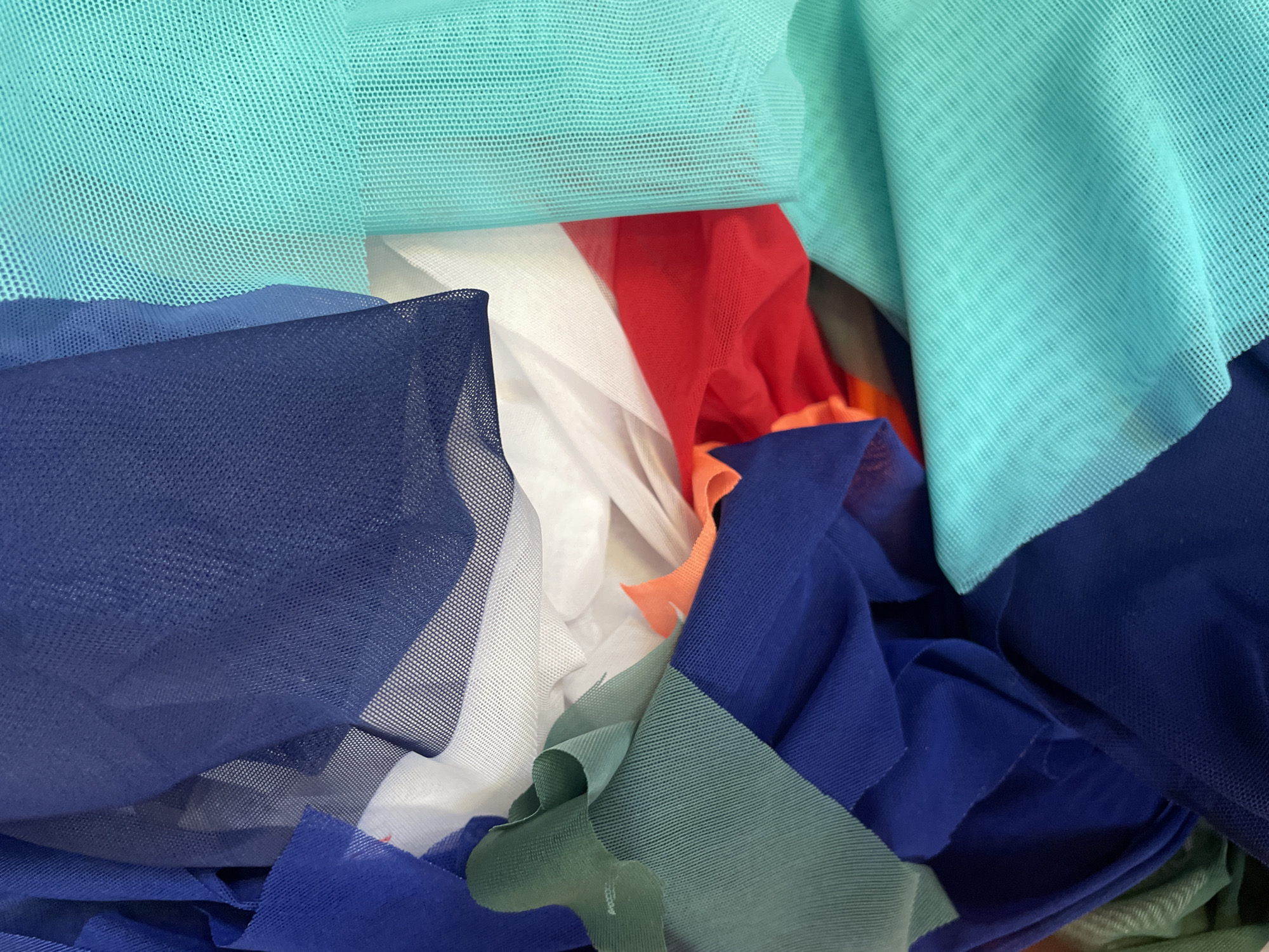 The image shows a pile of brightly colored fabric, primarily in shades of blue, green, red, and white. The fabrics appear to be made of a sheer, mesh-like material, possibly tulle or netting, and are loosely draped and folded. There are no people or discernible actions depicted. The environment is not visible beyond the arrangement of the fabric itself. The location in Sinyavino, Russia, is not visually represented in the image.