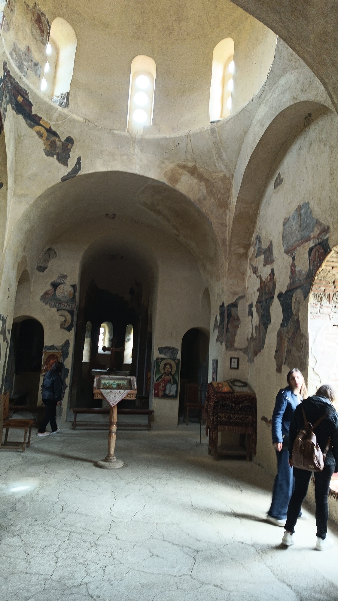 This indoor shot depicts a church interior in an unknown city. The architecture features arched ceilings, dome-like structures, and tall, narrow windows allowing light to filter in. The walls are adorned with deteriorated murals and paintings. A cross stands in the center of the room with a table in front of it. There are two women in the foreground, with one standing with a blue jacket while the other has a black jacket and a brown backpack. A person can be seen at the doorway behind the cross. Additional religious artifacts and furniture are placed around the room. The floor is uneven and patterned with cracks.