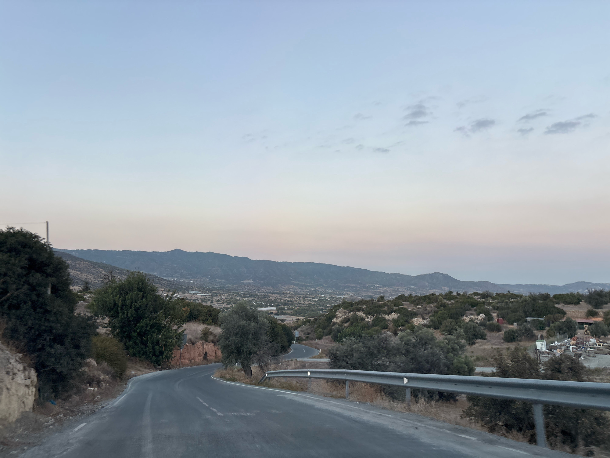 The image depicts a winding asphalt road in Armenochori Community, Cyprus. The road curves from the foreground into the mid-ground, bordered by dry grass, scrub brush, and scattered trees. A metal guardrail runs along the right side of the road. In the background, rolling hills covered in vegetation stretch towards a distant mountain range under a pale, dusky sky with scattered clouds. Several buildings and structures are visible in the valley between the road and the mountains, suggesting a rural or semi-rural settlement. The scene appears to be captured during either dawn or dusk.
