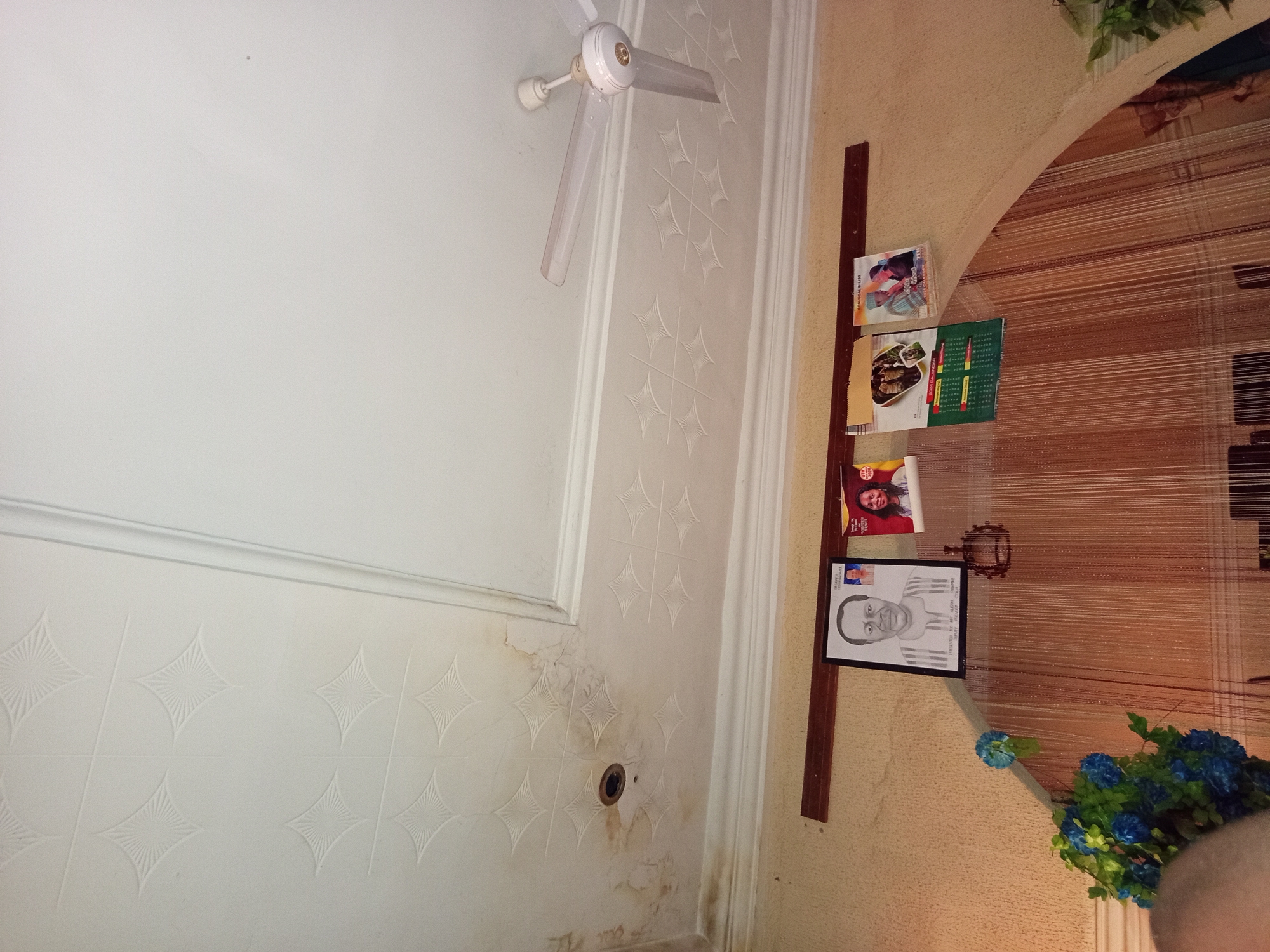 The image shows a room with a textured white ceiling, a ceiling fan, and a wall adorned with framed pictures and calendars. In the upper left of the ceiling, there are water stains. A ceiling fan with white blades is visible in the upper right corner. Below the ceiling, a dark wooden rail runs horizontally across the wall. Hanging on the rail are several items. On the left is a framed black and white portrait of a man in traditional clothing. To its right are three calendars, two with images of couples and one with an image of food. The wall below the rail is a textured yellow color. The lower portion of the image reveals a curtain with vertical strands. There is also a floral arrangement with blue flowers on the left side of the wall.