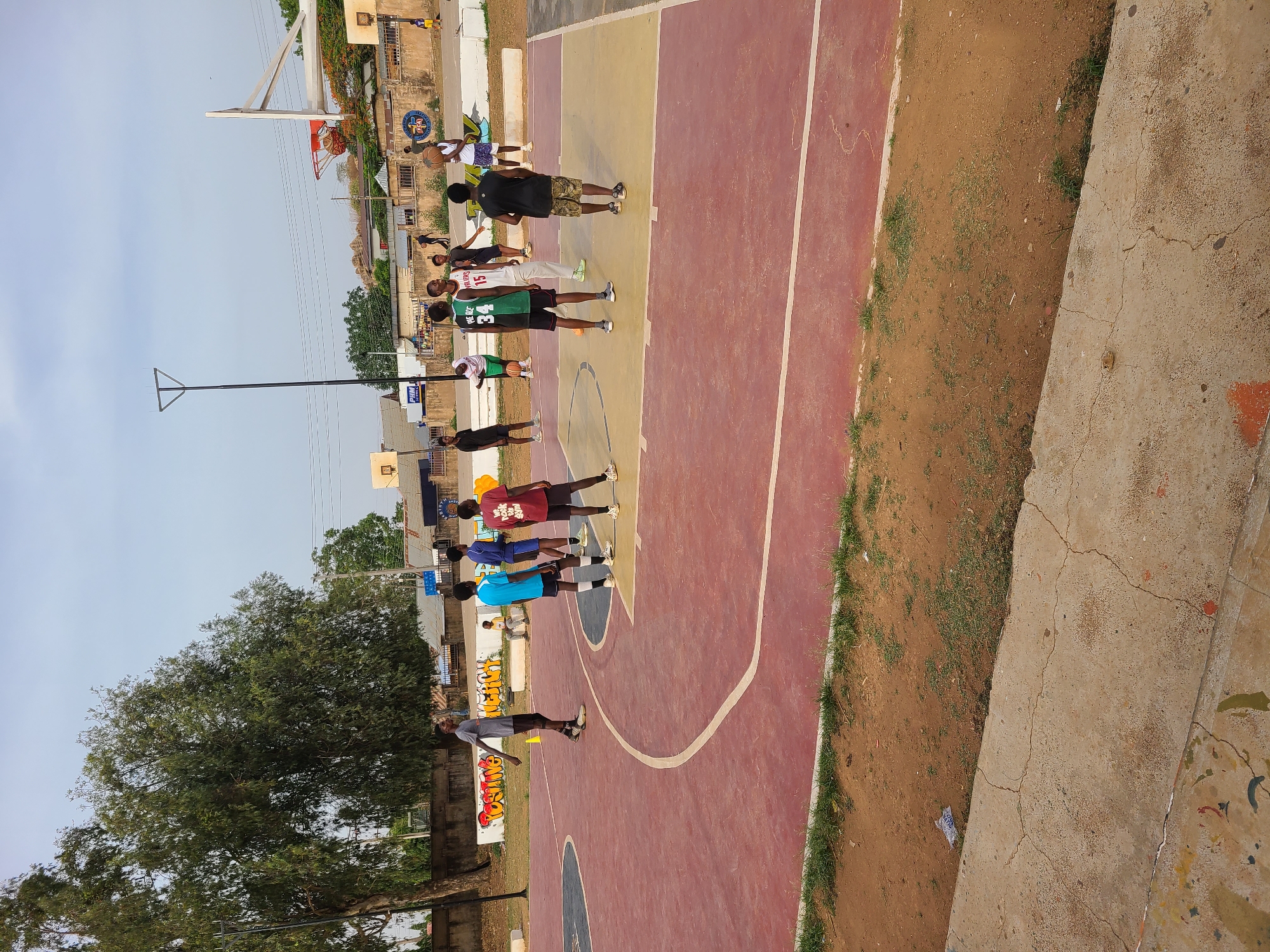 The image captures an outdoor basketball court scene in Jos, Nigeria, under a bright, clear sky. The court itself is distinctive with alternating red and yellow painted sections, marked with white lines, showing signs of use. A main basketball hoop with a white backboard and a red net is visible on the left side of the frame, with a smaller, portable hoop in the distance.
Approximately ten young males, varying in age from pre-teen to young adult, are present on the court. They are dressed in casual attire, including t-shirts and shorts, with some wearing basketball jerseys. Notable text on jerseys includes "WARRIORS 15" and "HE RO 34", and the name "AKO" is visible on another player's shirt. An adult male in a black t-shirt and camouflage shorts stands centrally, while others are distributed across the court, some holding basketballs. The overall activity suggests a casual basketball practice session or a pickup game.
In the background, various buildings, utility poles, and power lines indicate an urban or semi-urban setting. A large, verdant tree occupies the lower left corner of the frame. The court is bordered by a strip of dry earth with patches of grass, and concrete pathways or barriers. Graffiti or painted text, including "WELCOME" and "AKO", is visible on a wall or barrier adjacent to the court. A partial KFC logo can also be spotted on a building in the background. The scene depicts a lively daytime recreational gathering.
