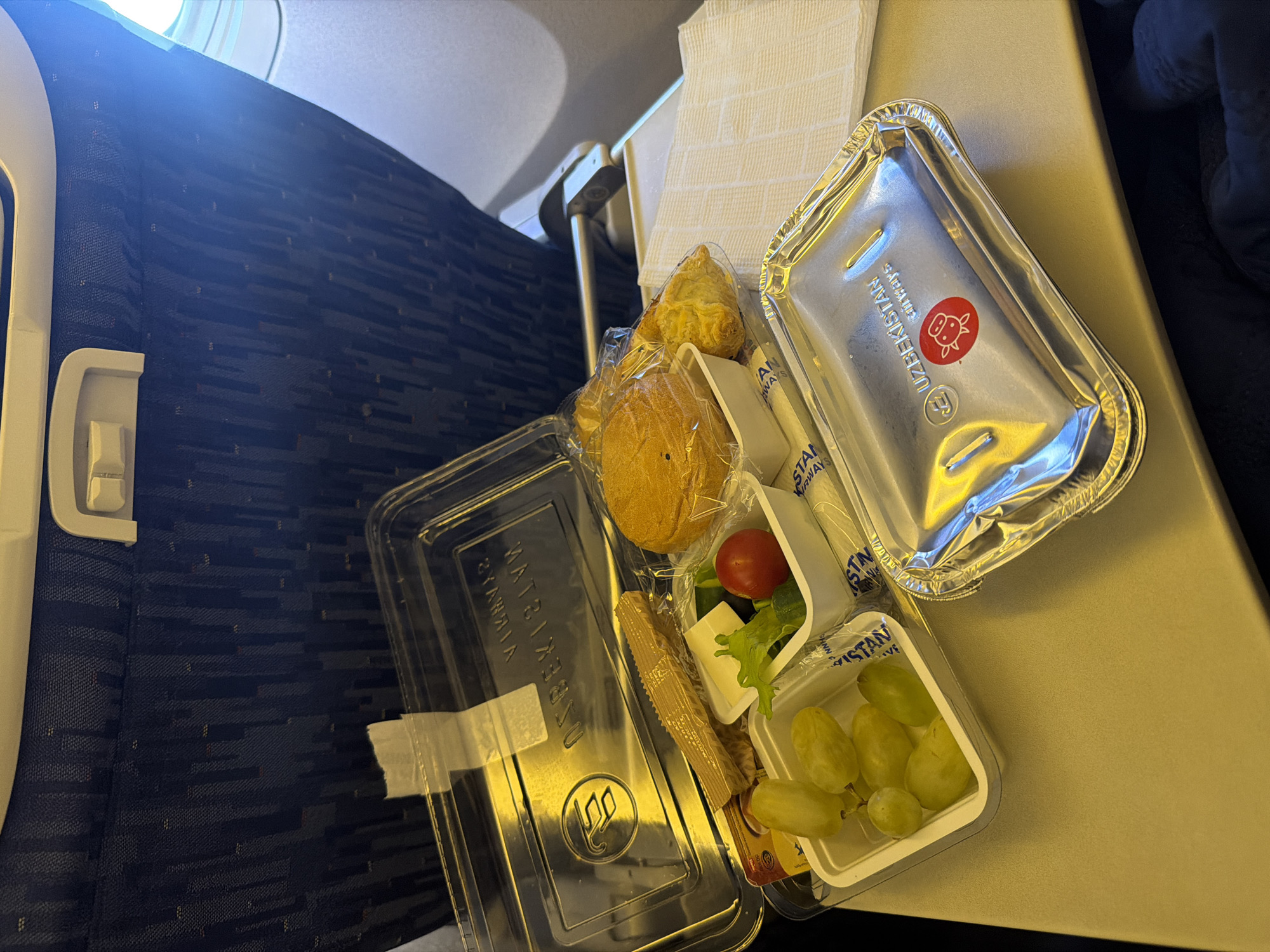An overhead shot shows an airplane meal on a tray table. The meal is served in a clear plastic container with a lid that has been partially opened. Inside, there is a bun, a pastry, grapes, cheese, a cherry tomato, and a small salad. A foil-covered dish is also present. The tray table is on the back of a seat, which is upholstered in blue fabric with a subtle pattern. The window to the right shows a bright, overcast sky, suggesting it is daytime. The overall lighting is warm, with reflections visible on the plastic and foil surfaces. There is no indication of the location being the city of Dubai, United Arab Emirates.