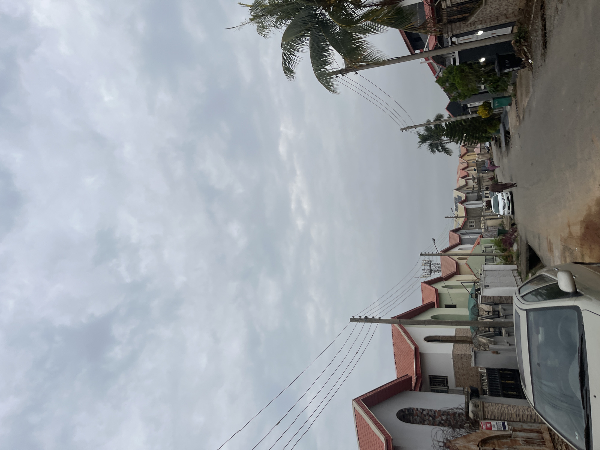 The image depicts a street scene in Abuja, Nigeria, characterized by a row of modern residential houses with red tiled roofs. The sky is overcast with thick, gray clouds. Electrical poles and overhead wires are visible along the street, typical of urban infrastructure.
In the foreground, a silver car is parked on the left side of the road, partially obscuring the view of the houses. Further down the street, other cars, including a white sedan, are present. Several individuals are walking along the street, indicating pedestrian activity. The houses themselves appear to be well-maintained, with various architectural styles and fencing. Palm trees and other vegetation are scattered throughout the scene, adding to the tropical urban landscape. The road surface appears to be paved. The overall impression is that of a residential neighborhood in a Nigerian city on a cloudy day.