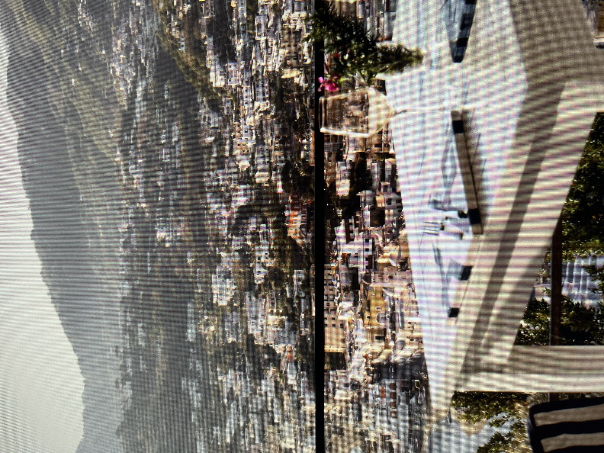 The image shows a scenic view of a town built on a hillside, with many white and yellow buildings cascading down towards a body of water. In the foreground, a white table is set for a meal, with a glass of white wine, cutlery, and a small floral arrangement. The overall impression is one of a beautiful, sun-drenched location, possibly a coastal town. The scene does not depict any elements that would suggest the city of Łódź, Poland.