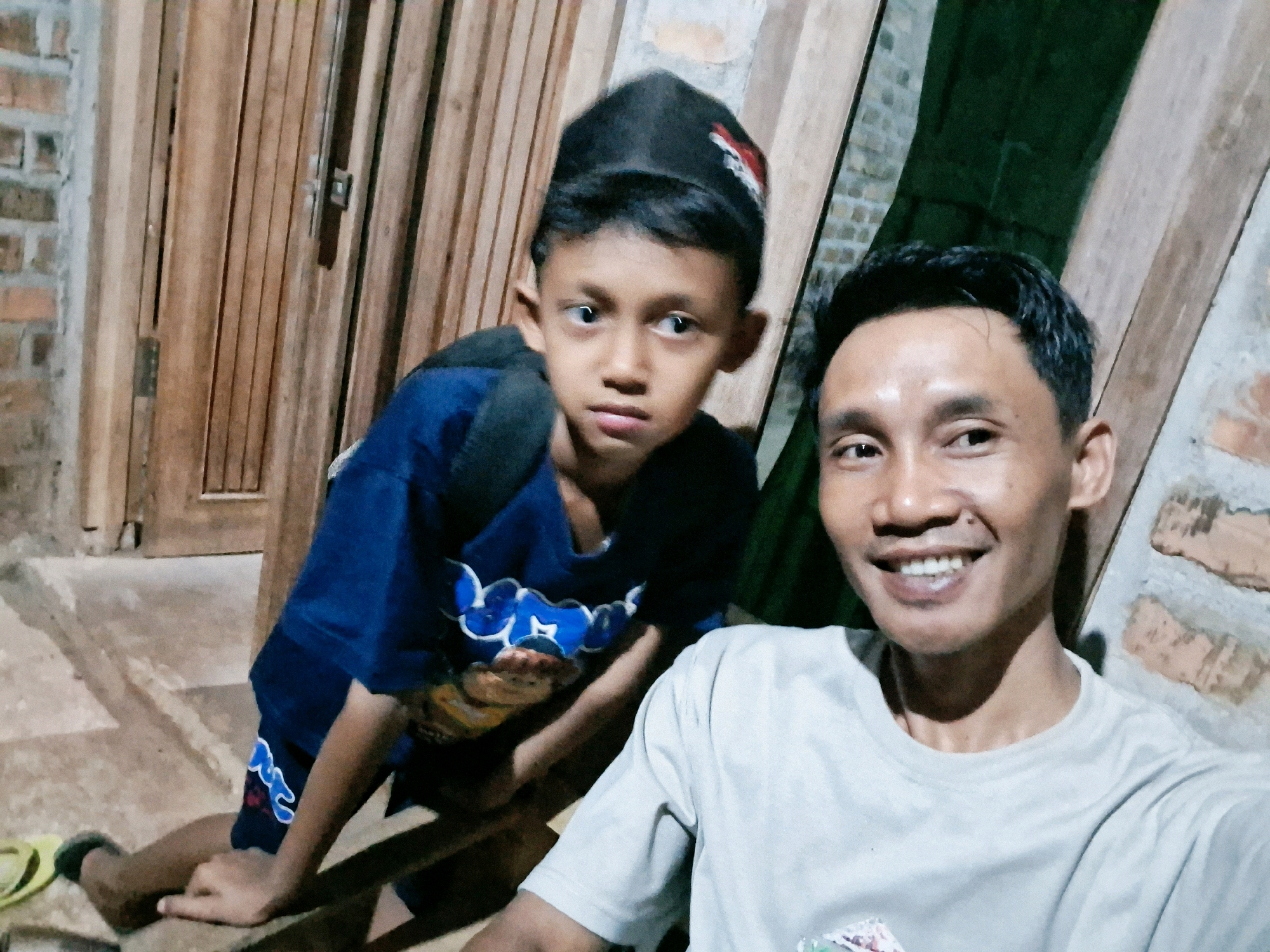 A medium shot shows two males, likely father and son, posing for a selfie in what appears to be an outdoor setting in Karang Sari, Indonesia. The younger male, on the left, is wearing a blue shirt with a white graphic design and a black cap. He is leaning on a wooden surface. The older male, on the right, is smiling widely and wearing a light-colored t-shirt. Behind them, a wooden door and brick wall are visible. The ground is concrete.