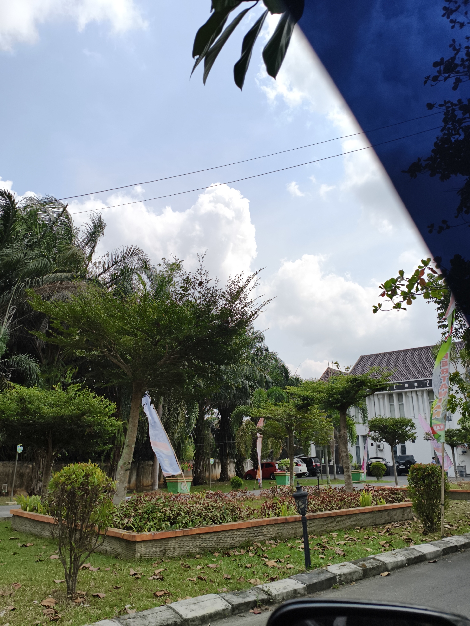 The image depicts a street view in Kemelak Bindung Langit, Indonesia, on a bright, cloudy day. In the foreground, a paved road with a concrete curb is visible. Beyond the road, a landscaped area features a raised garden bed filled with low-lying plants and some fallen leaves on the grass. Several trees are present, including palm trees and a larger deciduous tree with a white flag draped around its trunk.
In the mid-ground, a white building with a dark tiled roof and columns can be seen. Several cars are parked in front of the building. Flags are displayed on poles in front of the building and in the landscaped area. The sky is partly cloudy with large white cumulus clouds against a light blue backdrop. The upper right portion of the image is partially obscured by a dark blue, possibly reflective surface, with some green foliage visible against it. No people are clearly identifiable in the scene.