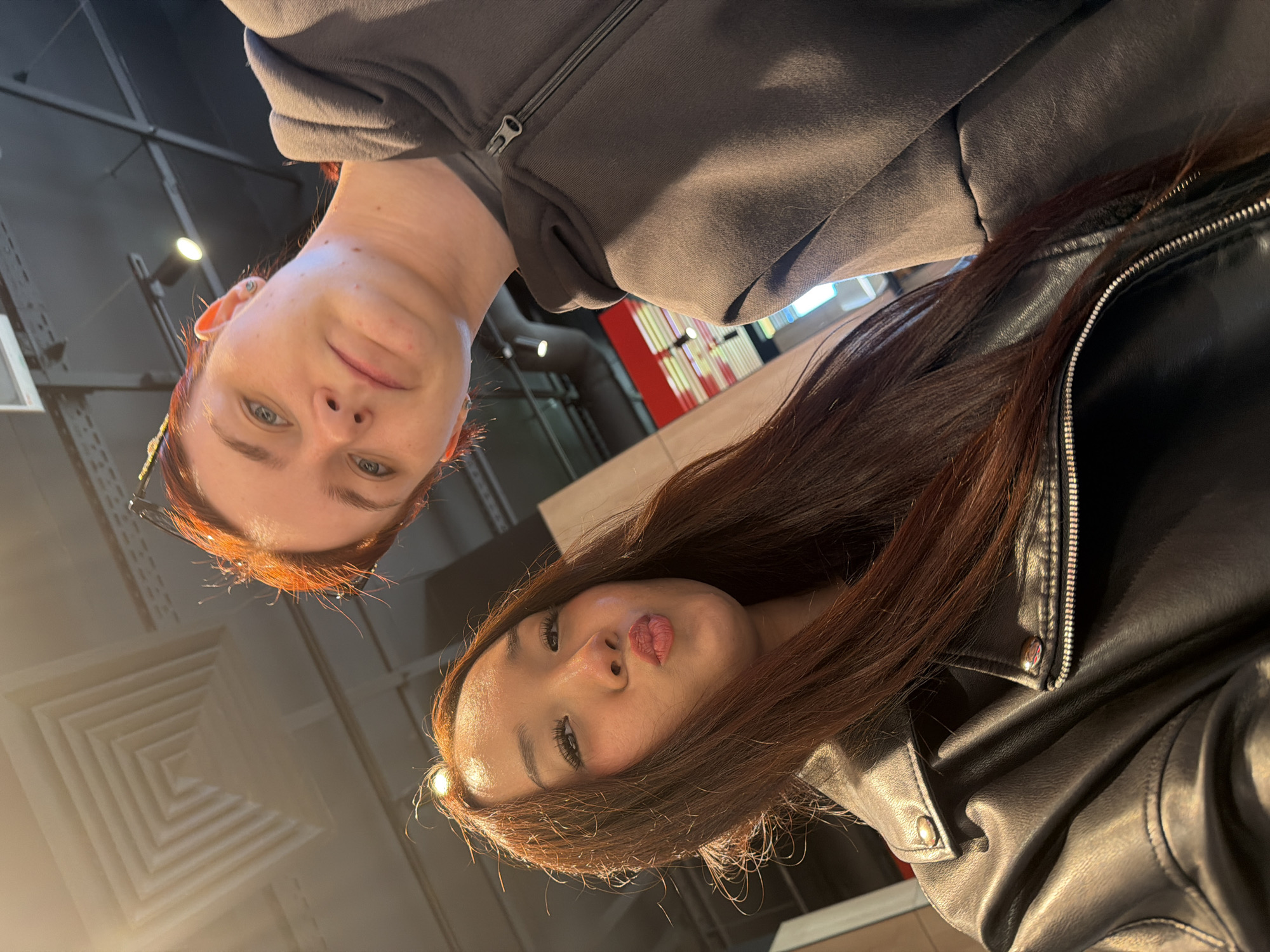 Two young women are taking a selfie indoors. The woman on the left has long, dark auburn hair and is wearing a black leather jacket. She is puckering her lips for the photo. The woman on the right has shorter, brighter red hair and is wearing a dark gray hooded sweatshirt. She is looking towards the camera with a neutral expression. The background suggests an indoor commercial space with a high ceiling and industrial lighting fixtures. The lighting is warm and bright, casting shadows. The scene is likely a casual setting, such as a cafe or a store, in Saint Petersburg, Russia, given the provided context.